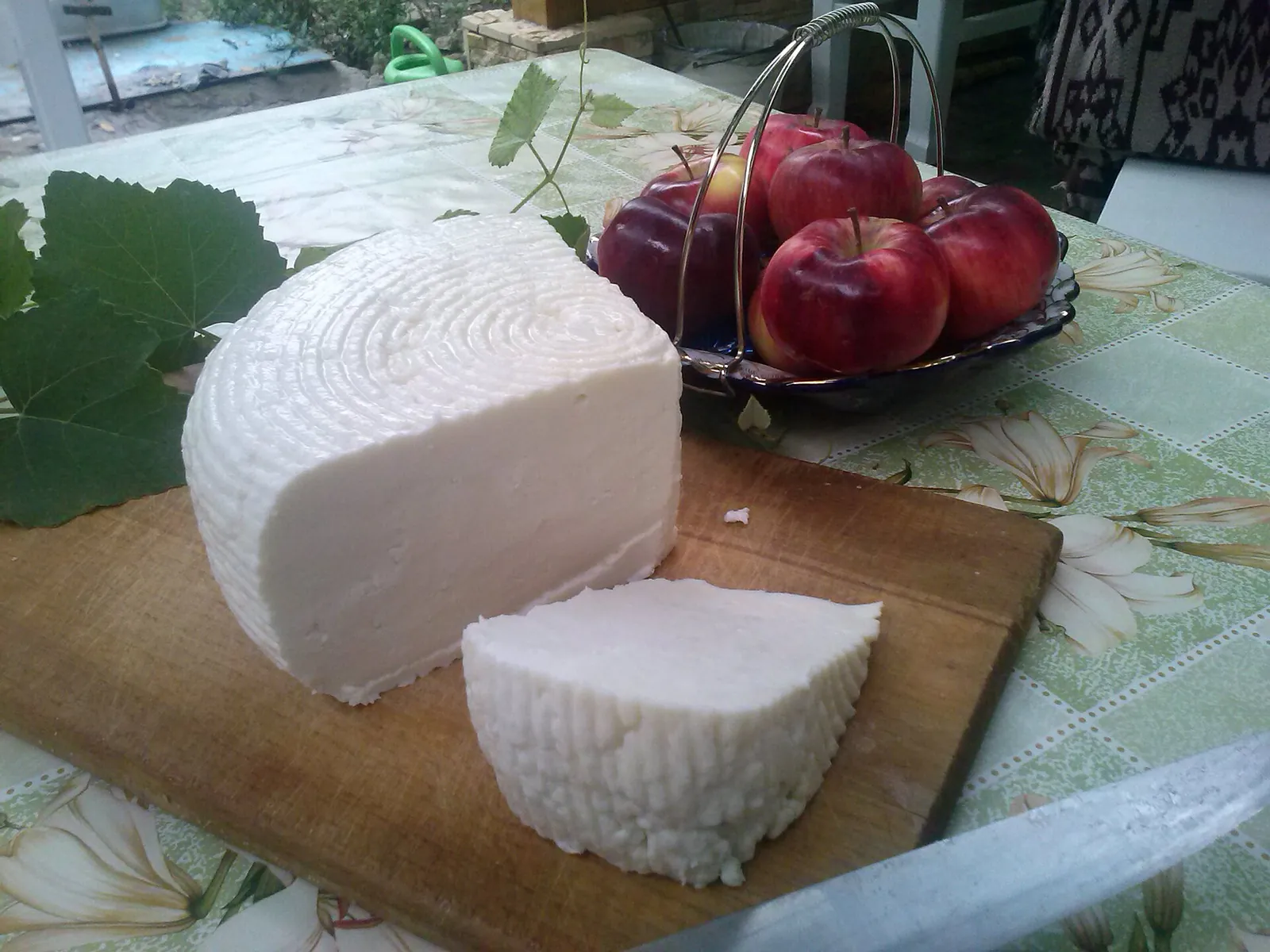 Adyghe cheese wheel and a cut wedge on a wooden board beside a basket of apples and grape leaves