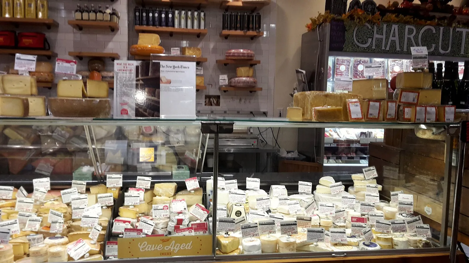 A long glass case in an American cheese shop holding rows of small cheese wedges with price tags, with charcuterie and jars on shelves behind.