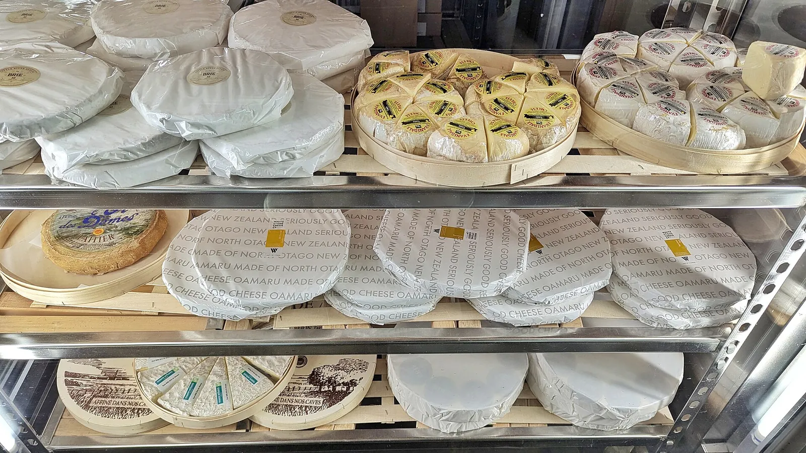 Shelves in a cheese shop in Wellington, New Zealand, holding soft cheese rounds wrapped in white paper and wooden boxes of cut wedges.