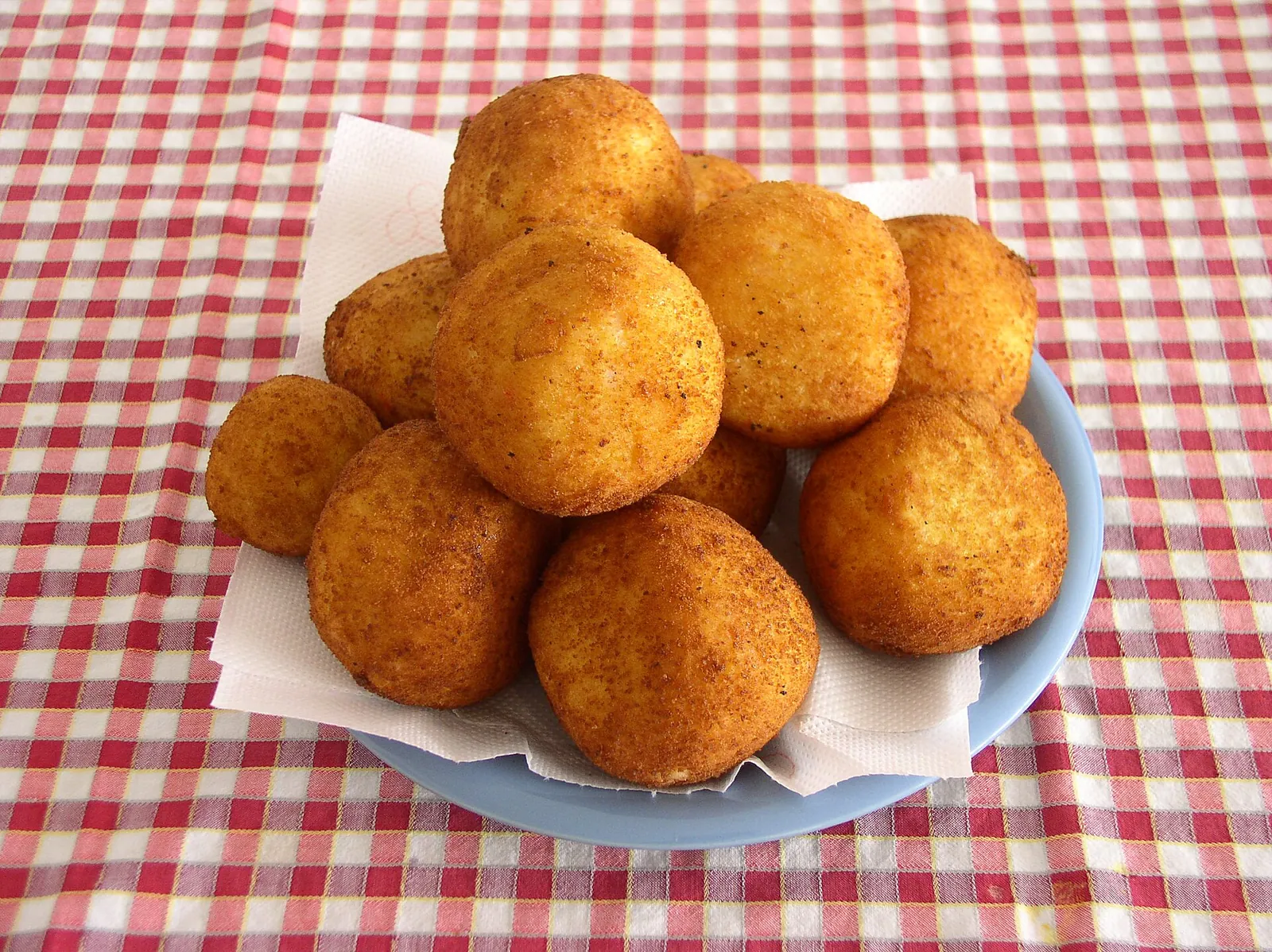 A pile of golden fried arancini rice balls in a blue bowl lined with paper, set on a red and white gingham tablecloth.