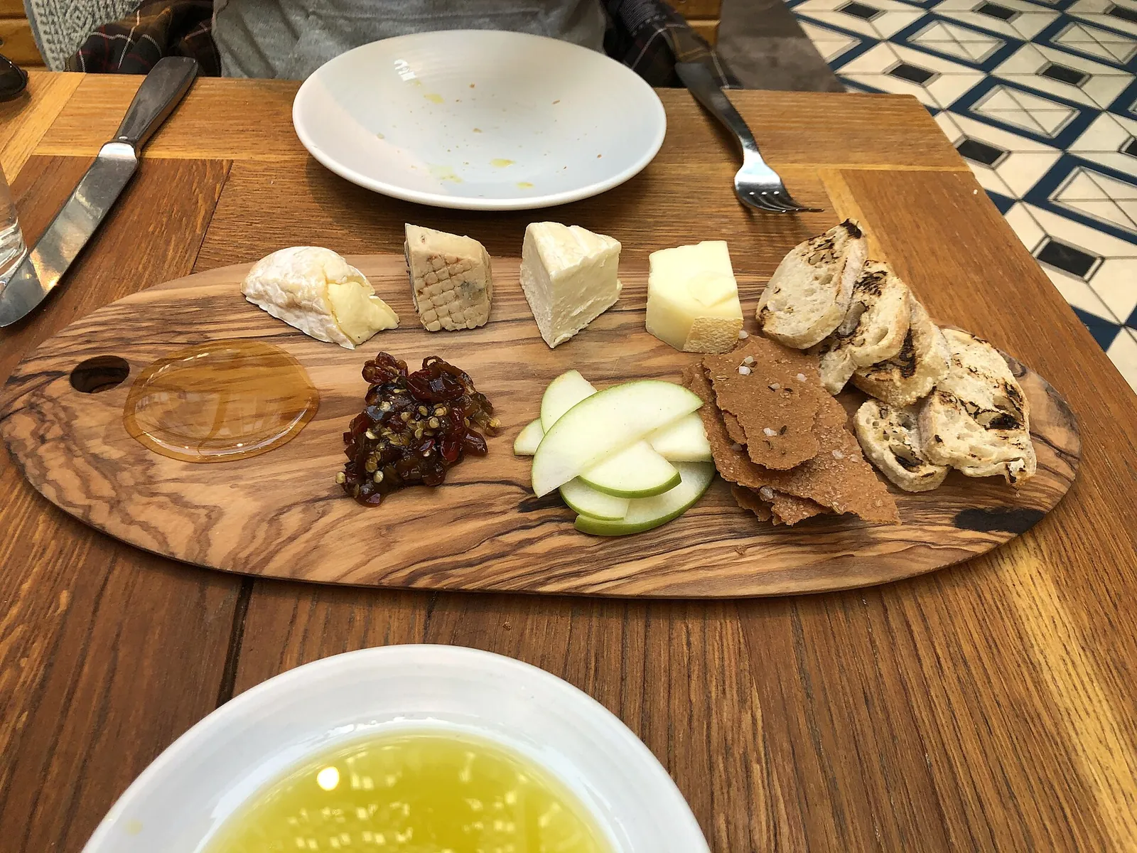 An olive wood board with small pieces of cheese, apple slices, fruit chutney and crackers beside a dish of olive oil.