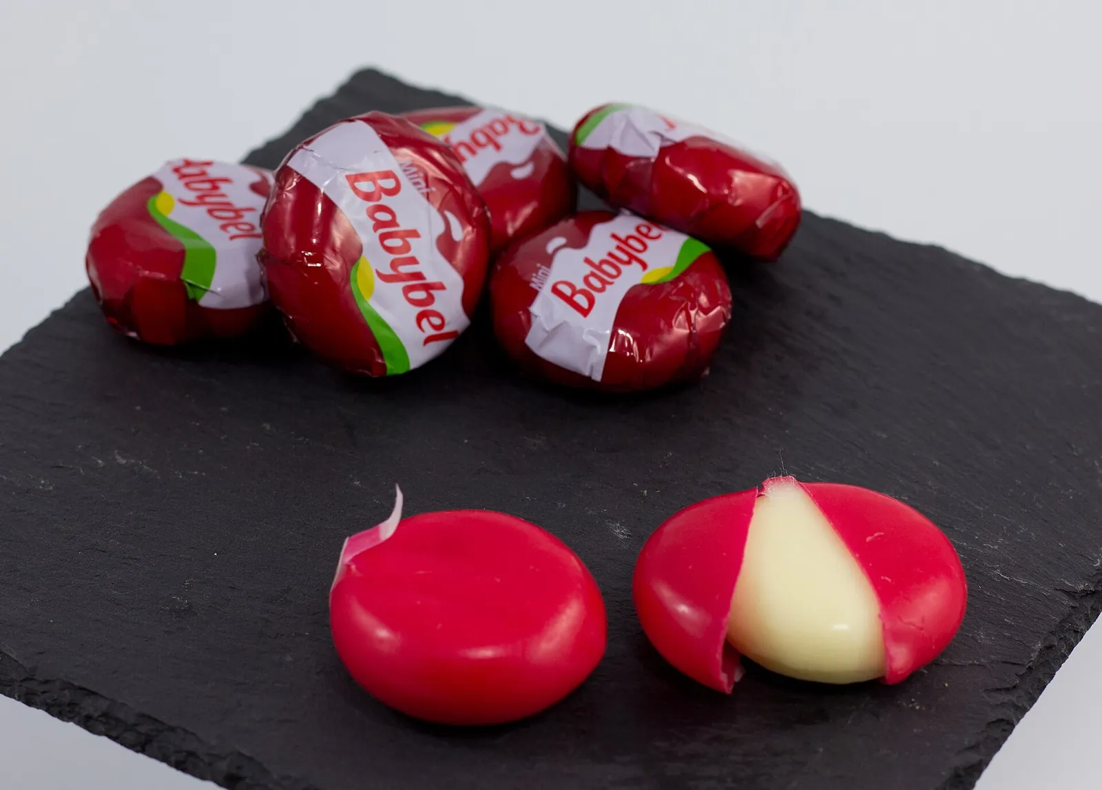 Mini Babybel cheeses in red wax and net wrappers on slate, two unwrapped and one cut open