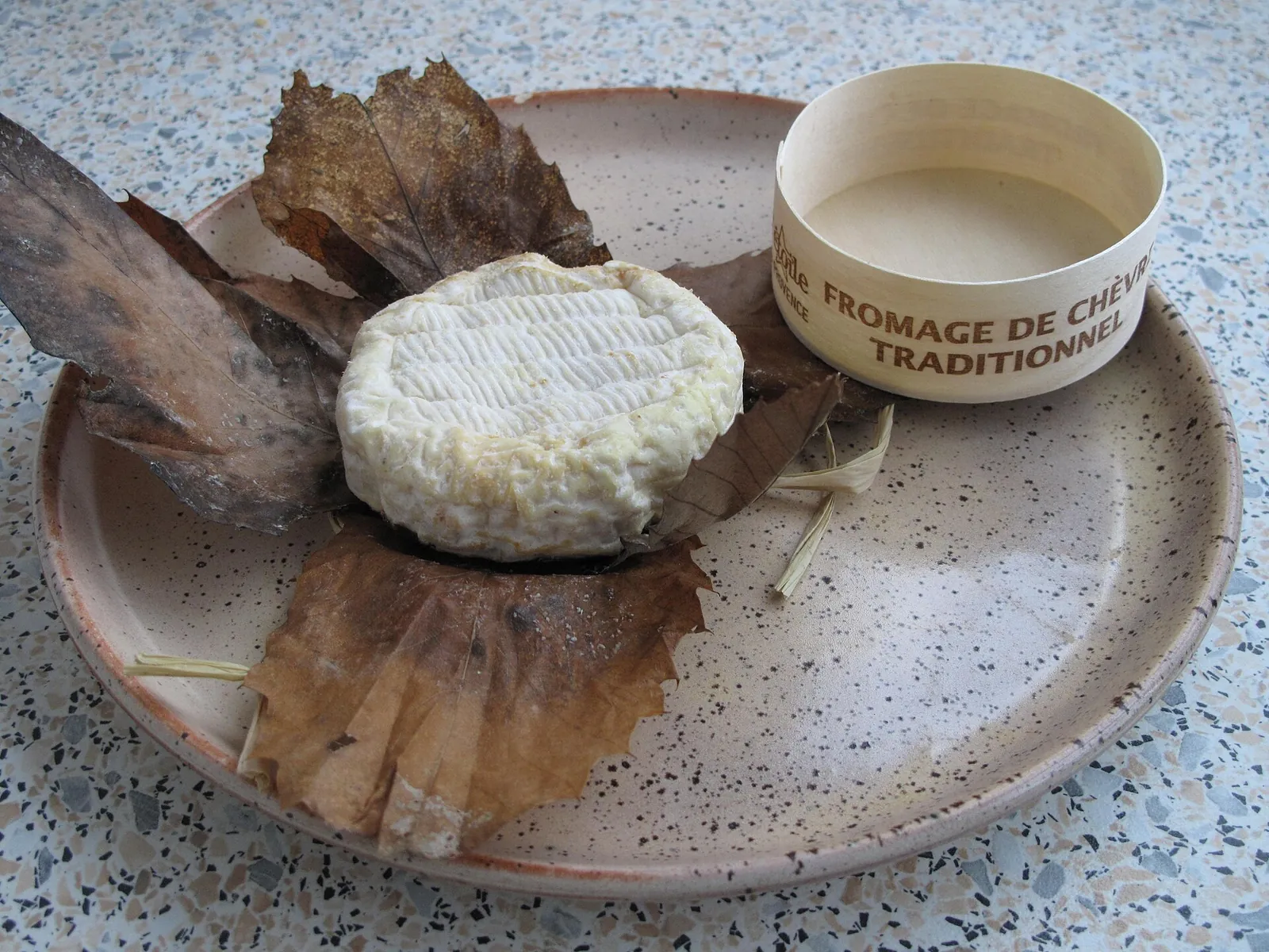 A round Banon goat cheese unwrapped beside its brown chestnut leaf wrapper and wooden box on a speckled plate