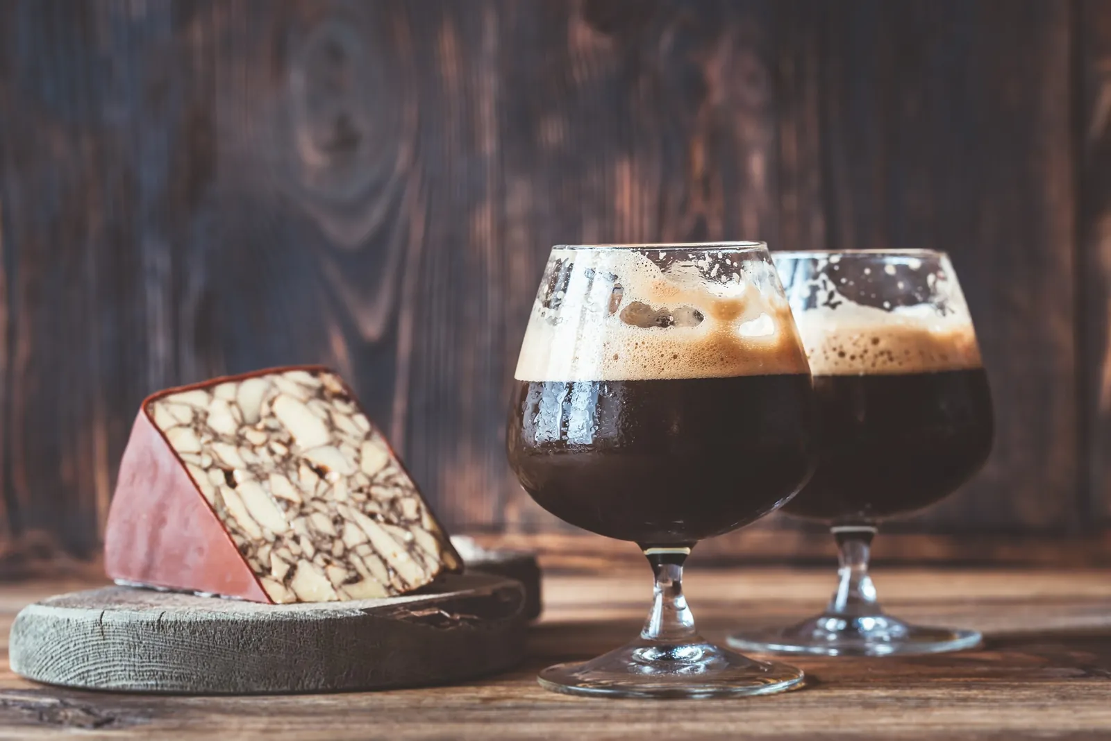 Two snifter glasses of dark stout with a foamy head beside a wedge of marbled porter cheddar with a brown waxed rind on a wooden board.