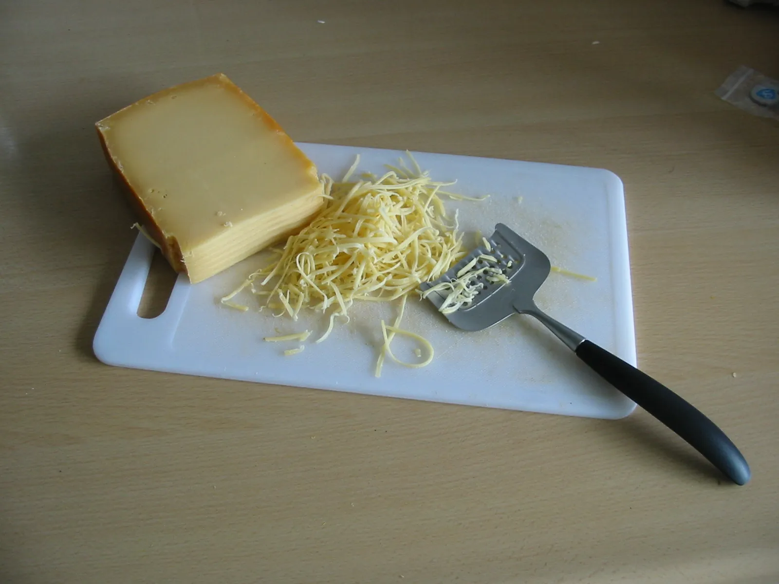 Block of semi hard cheese on a white cutting board beside a small pile of freshly grated cheese and a grater.