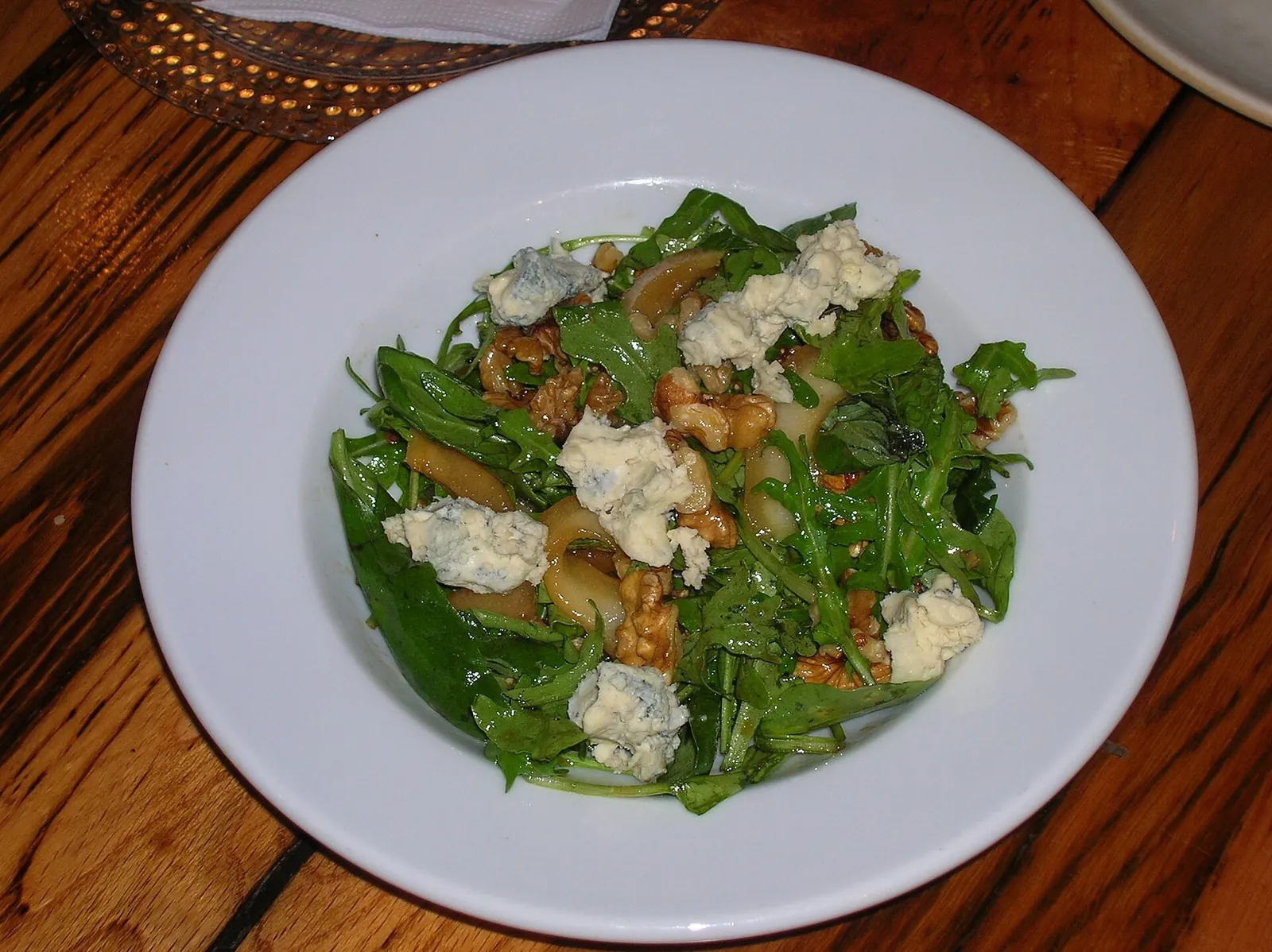 A white plate of arugula salad with crumbled blue cheese, walnuts and slices of grilled pear on a dark wooden table.