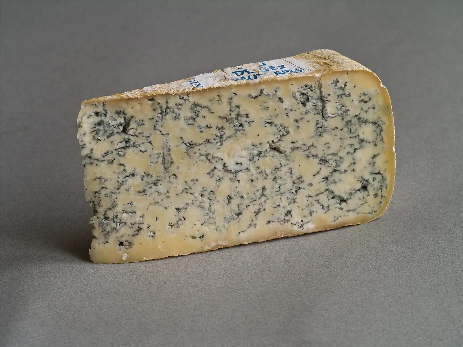 Triangular wedge of blue cheese with dense blue green veining and a natural rind on a gray background.
