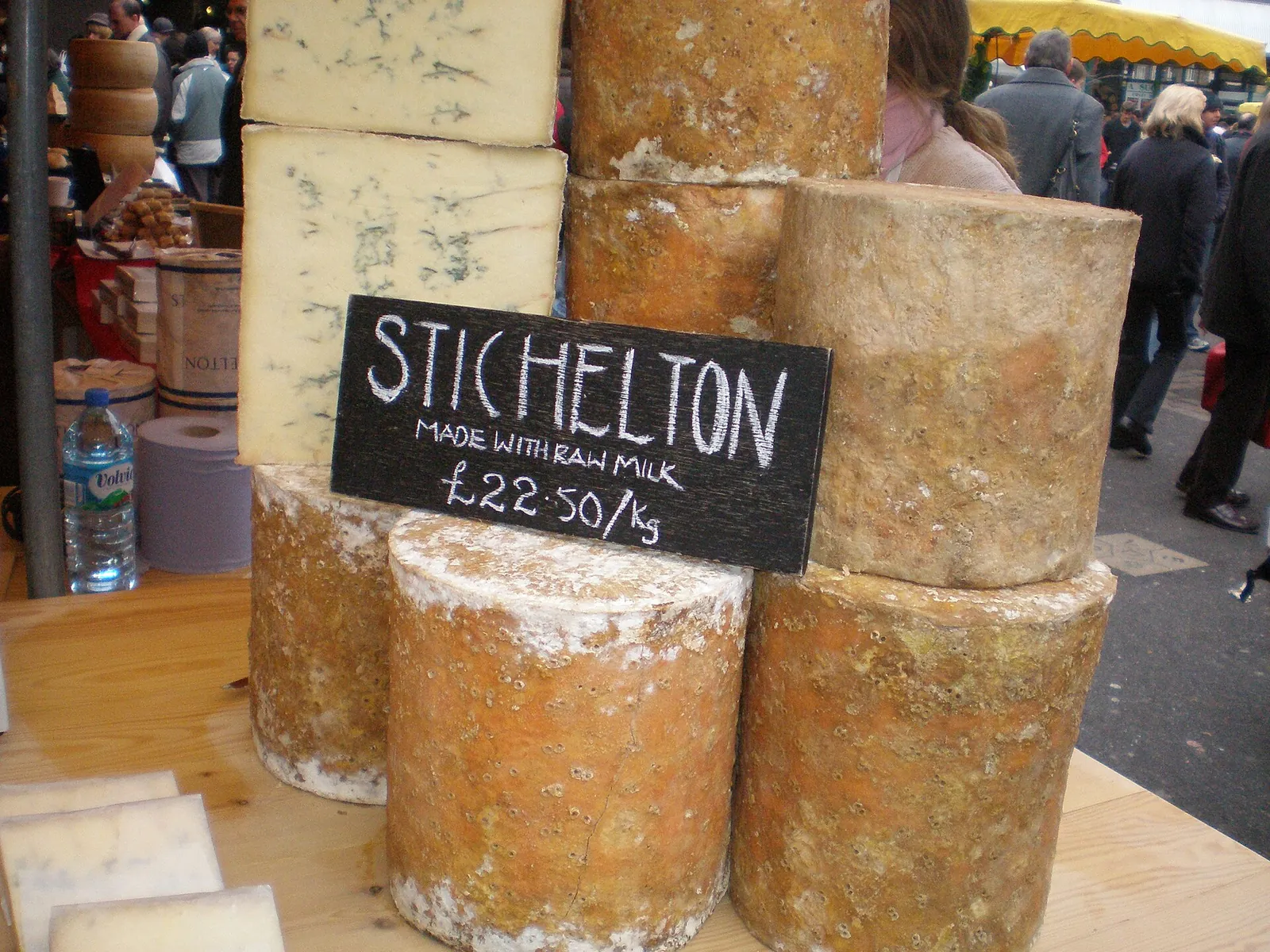 Tall cylinders of Stichelton, a raw milk English blue cheese, stacked on a market stall beside a chalkboard sign showing the price per kilogram.