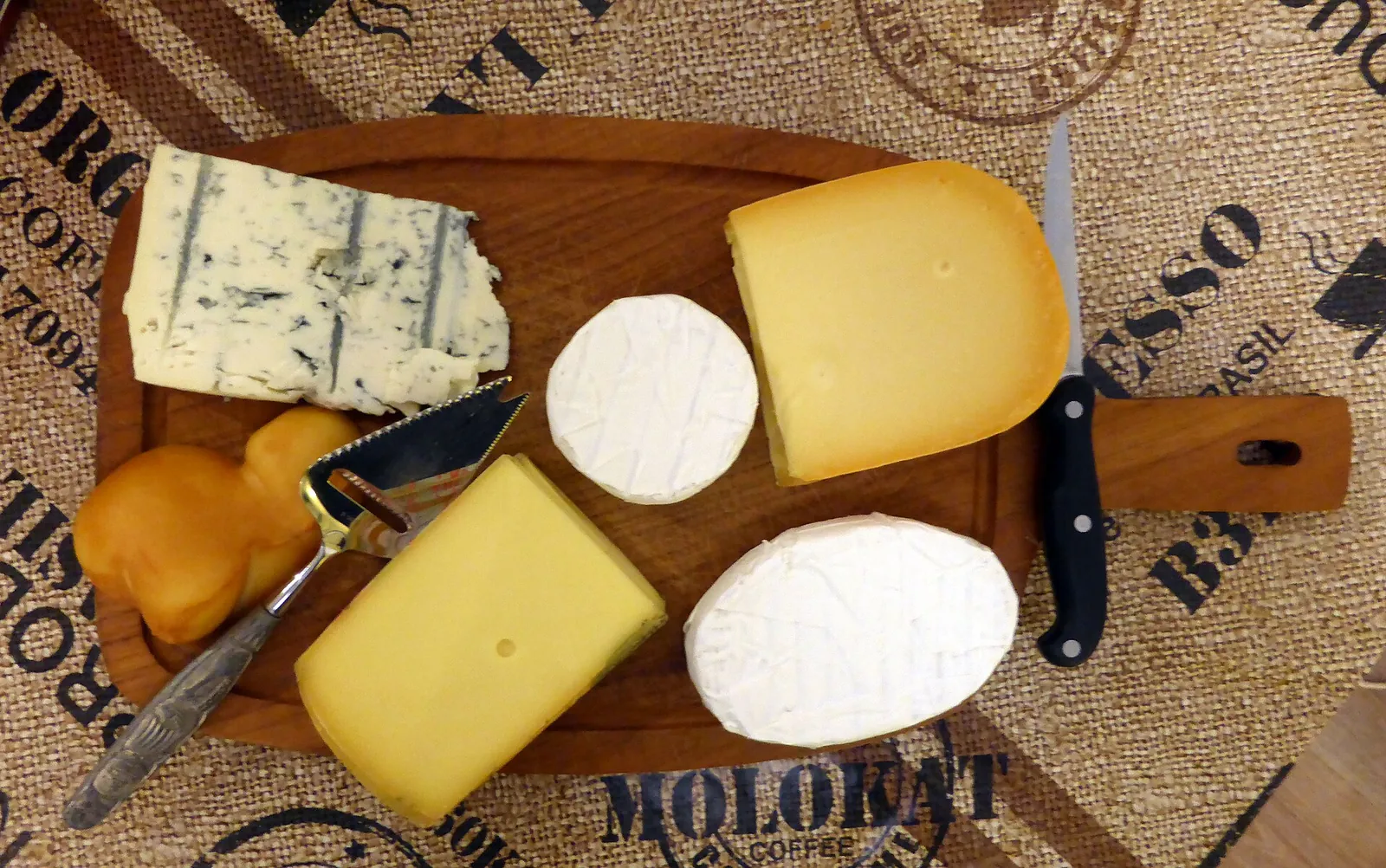 A wooden board with a blue cheese wedge, two white-rind rounds and two yellow Gouda-style wedges, with a cheese knife and a small cheese slicer
