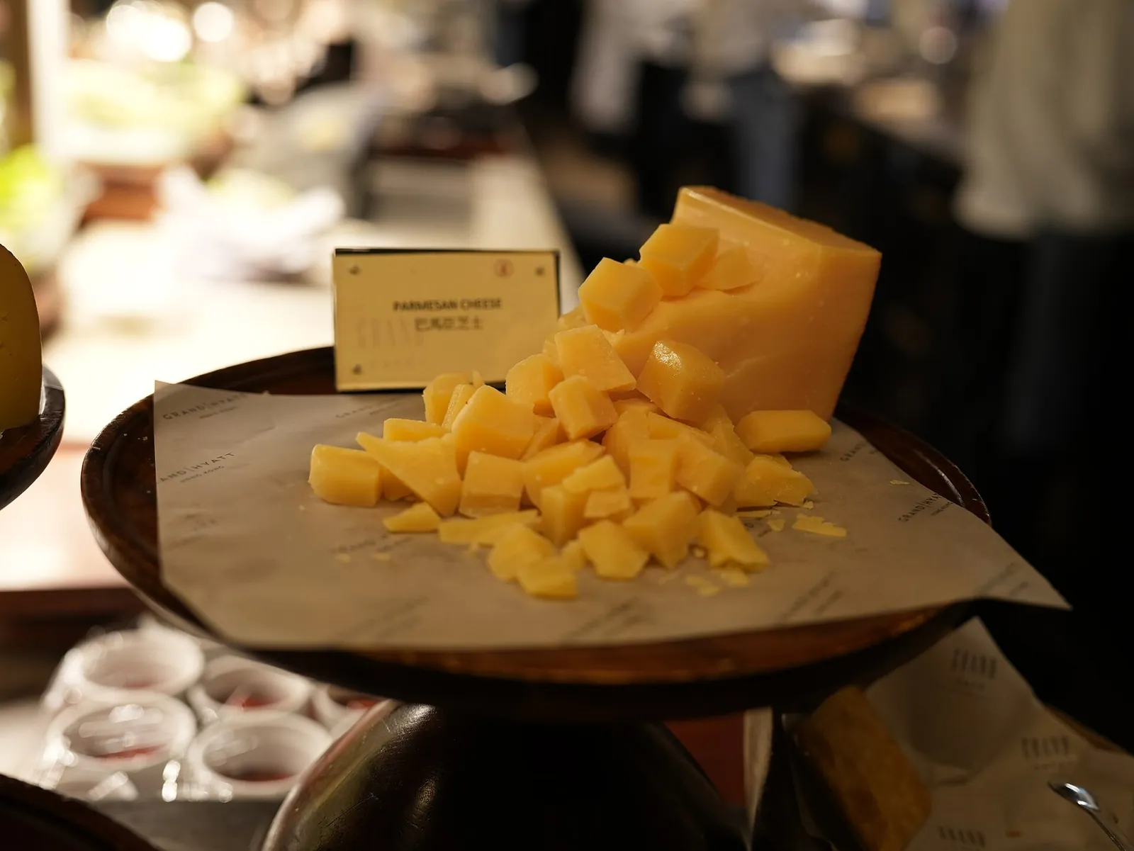Golden chunks of Parmigiano-Reggiano broken from a wheel and piled on paper on a stand, with a small sign beside them at a cafe
