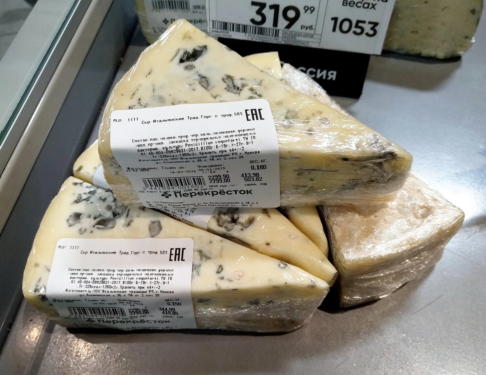 Three triangular wedges of blue cheese sealed in clear plastic wrap with printed labels, lying on a refrigerated shelf