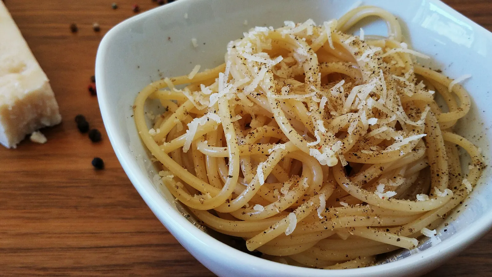 A white bowl of spaghetti tossed with grated cheese and cracked black pepper, with a wedge of pecorino and peppercorns beside it.