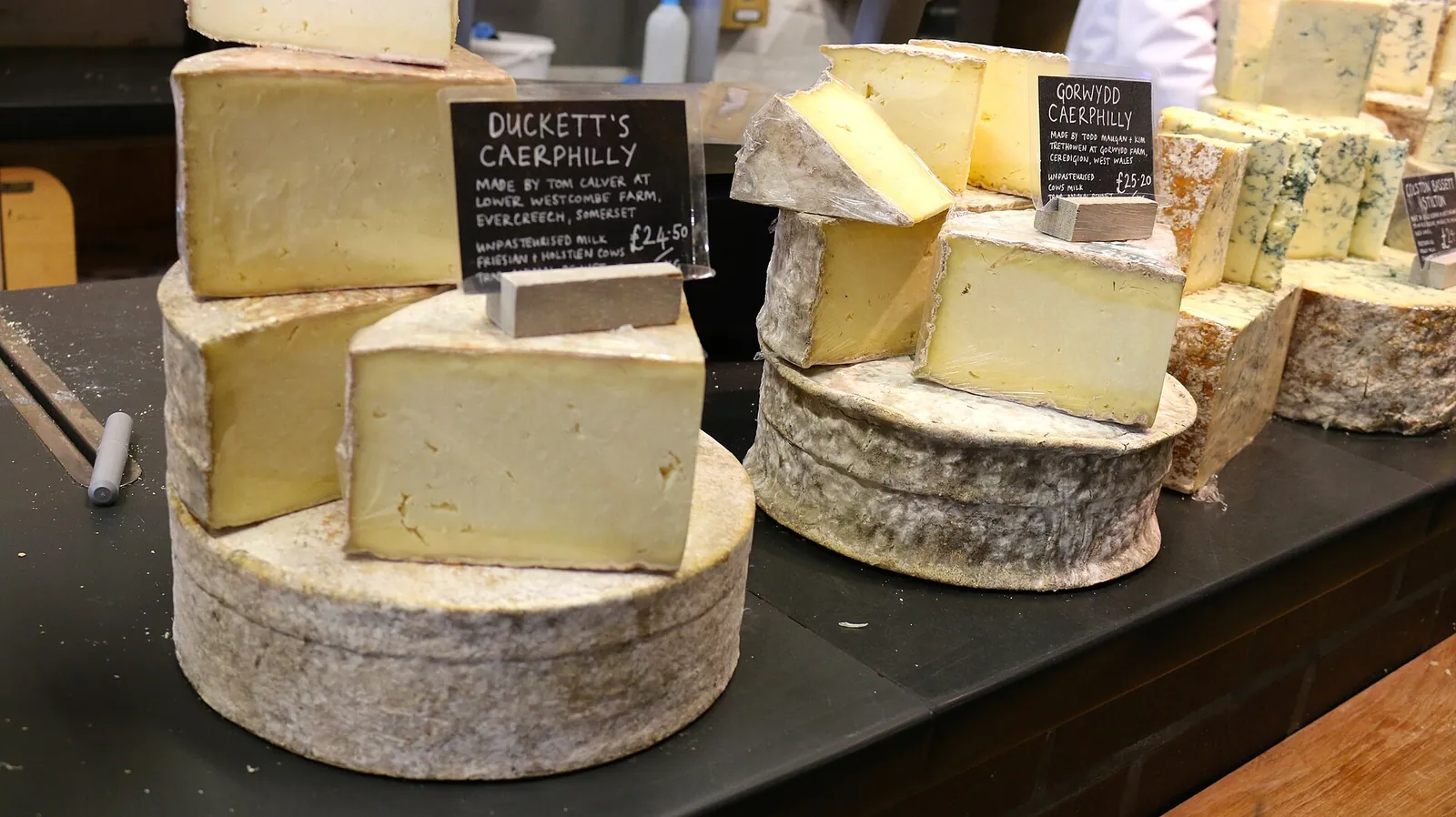 Wheels and cut pieces of Caerphilly at a cheese counter, with a handwritten sign for Duckett's Caerphilly