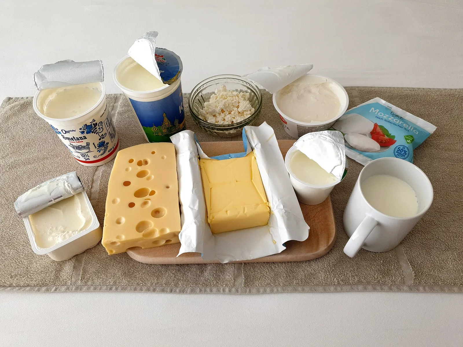 A table of dairy foods: Emmental and yellow cheese on a wooden board beside a glass of milk, yogurt cups and cream.