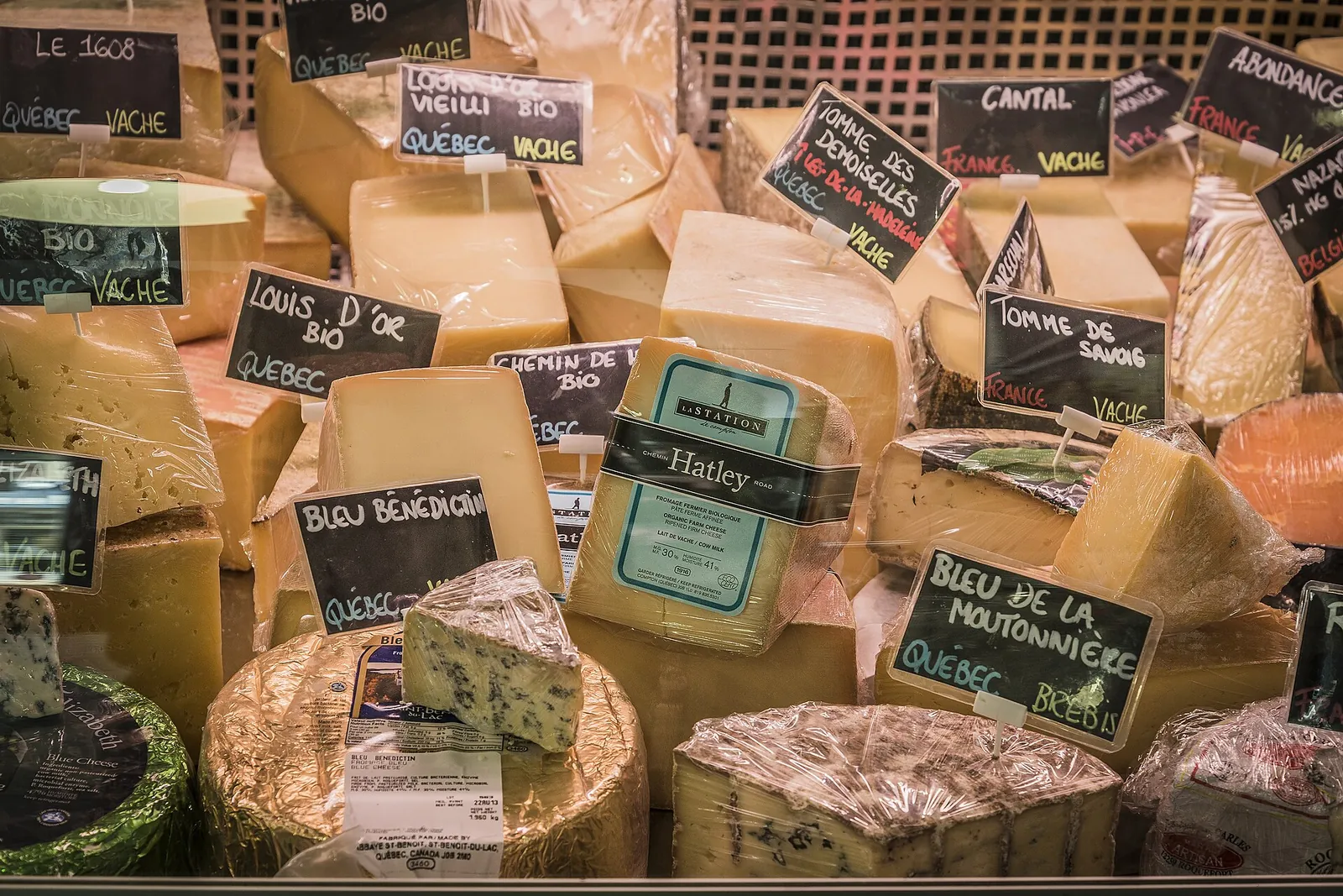 A crowded cheese counter in Montreal with wedges and wheels of Quebec cheeses, each marked with a chalkboard label naming the cheese.