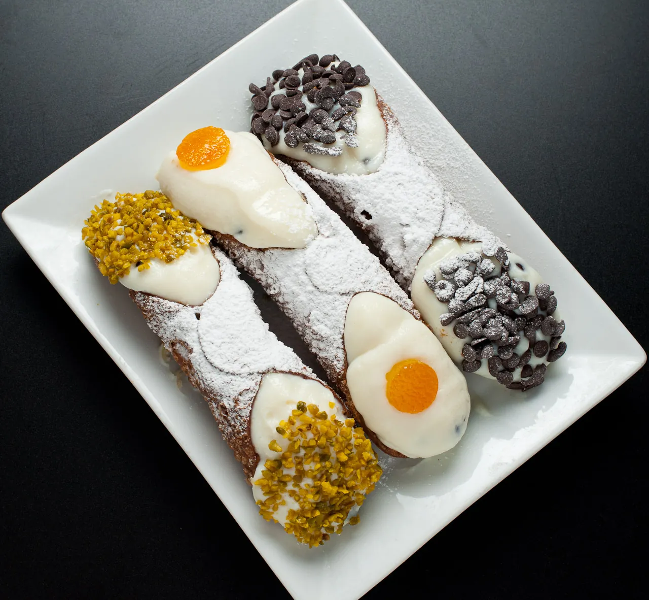 A cannolo with a crisp fried shell and a creamy white ricotta filling, dusted with powdered sugar and pistachio, on a patterned plate.