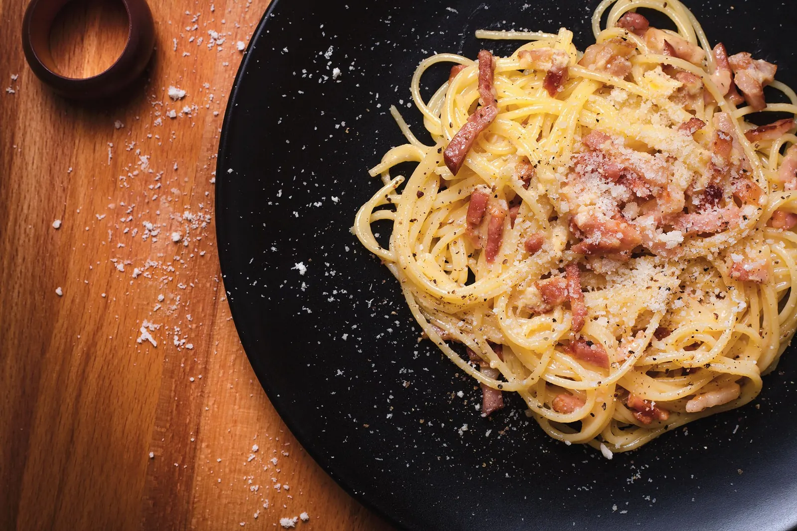Spaghetti carbonara on a dark plate, coated in a creamy sauce with crisp pieces of bacon and grated cheese scattered over the top.