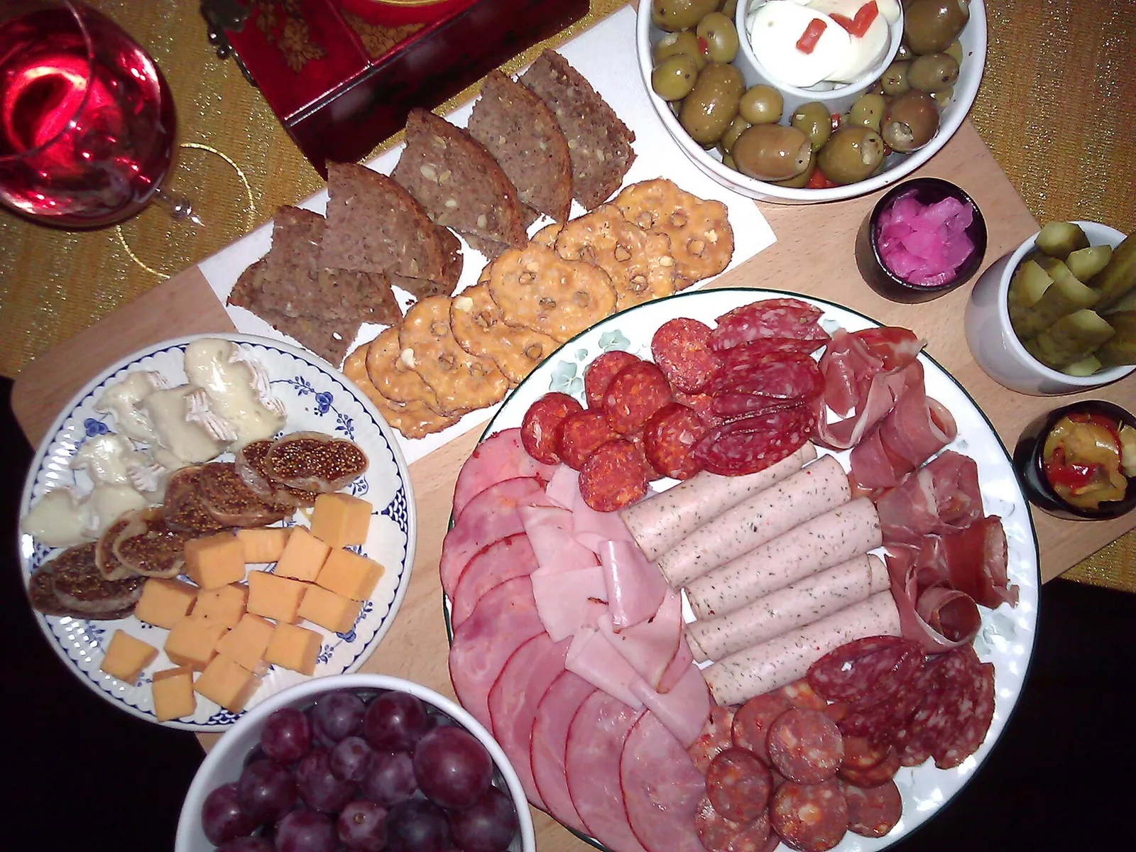 A tabletop spread seen from above with folded ham and salami, small plates of cheese cubes, crackers, bread, olives and grapes.