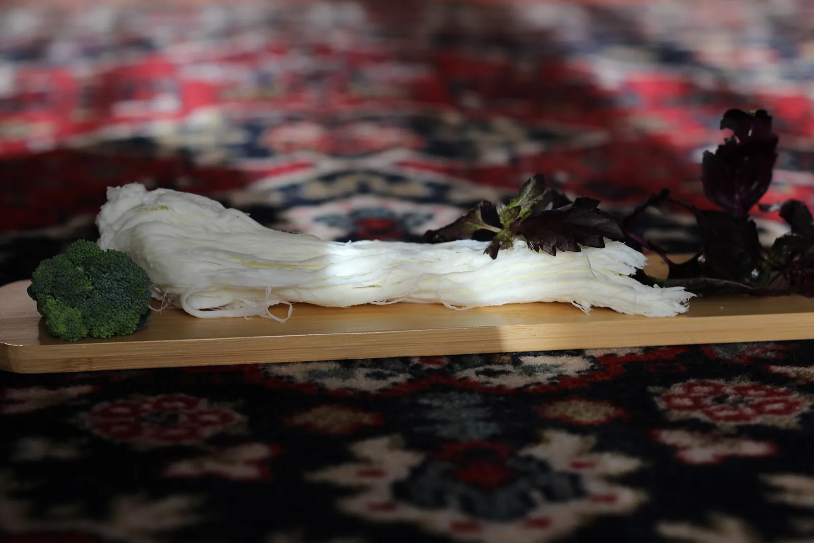 Strands of white chechil string cheese laid along a wooden board with broccoli and fresh herbs
