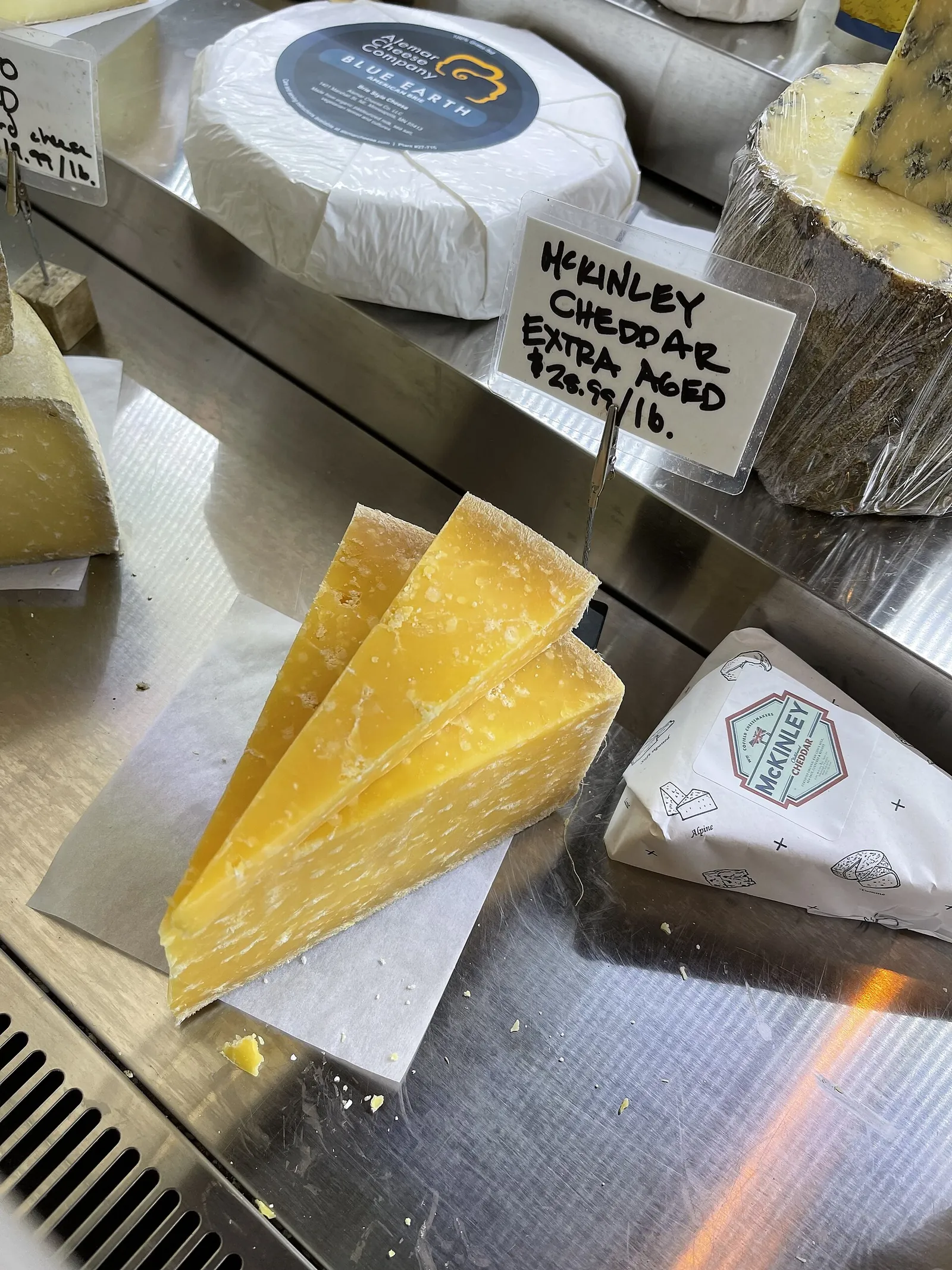 Wedges of extra-aged cheddar with a crumbly, crystalline texture at a cheese counter, under a sign reading McKinley Cheddar Extra Aged