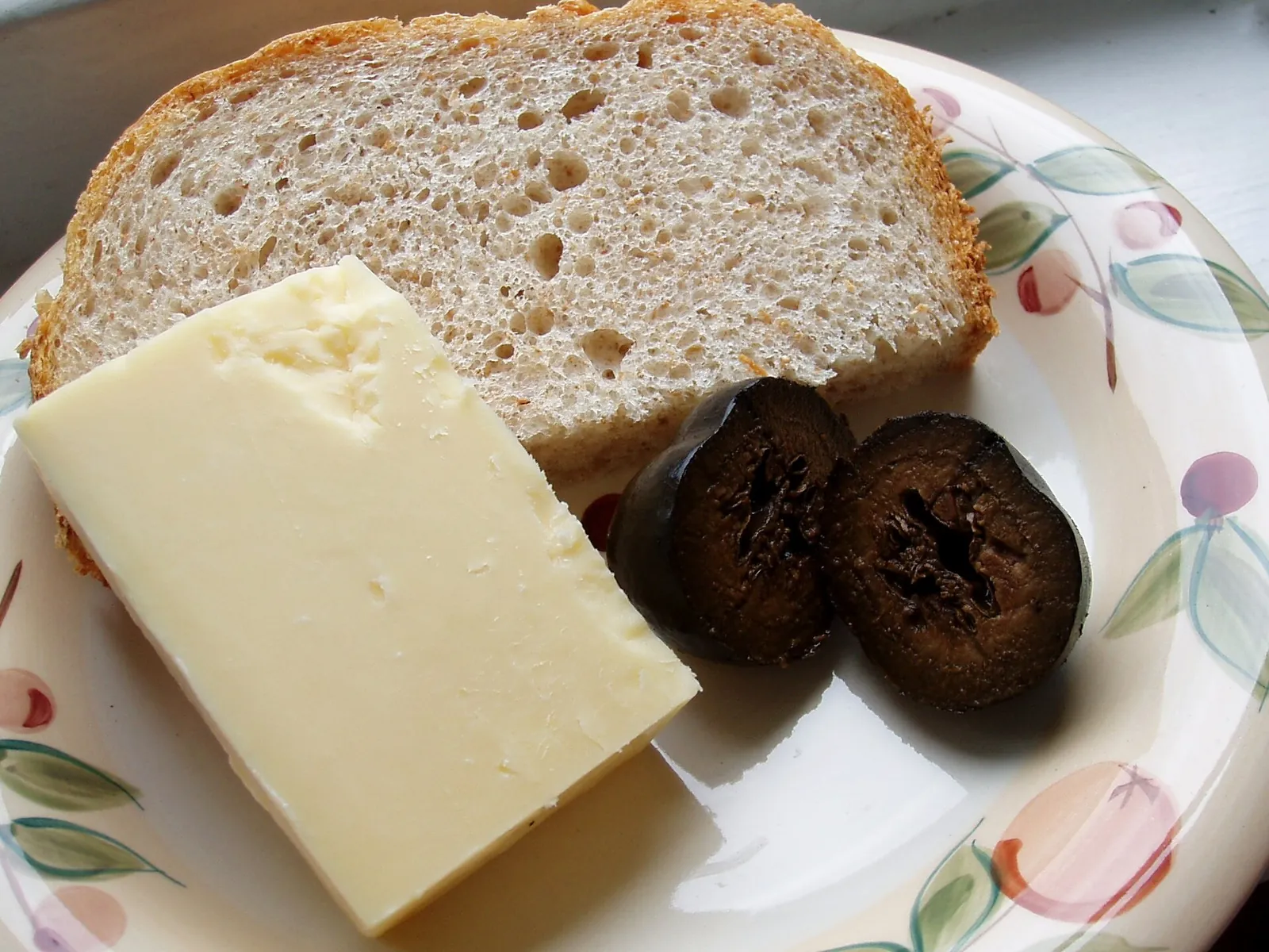 A thick slice of pale hard cheese on a flowered plate beside a slice of whole grain bread and pickled walnuts.
