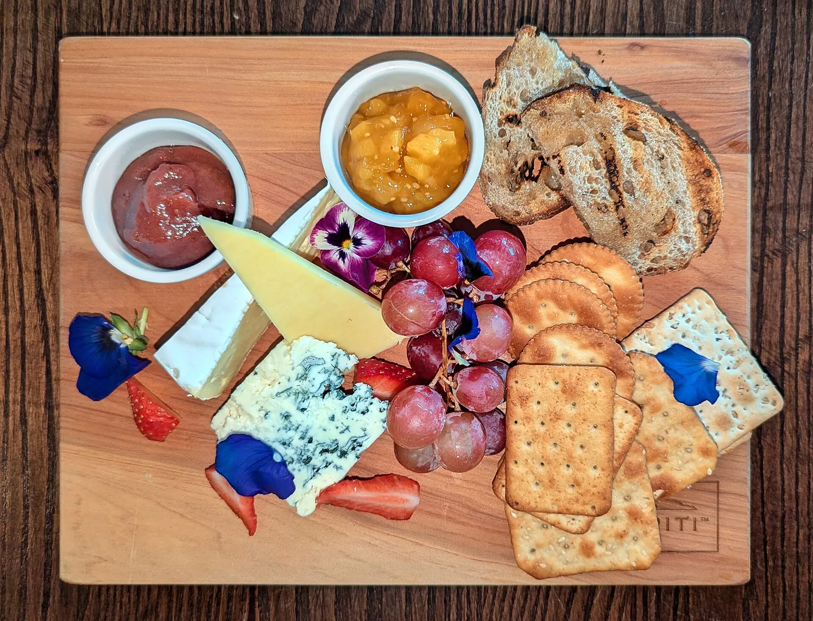 A wooden cheese board with blue and soft cheeses, crackers, grapes, strawberries, bread, edible flowers and two small dishes of chutney.