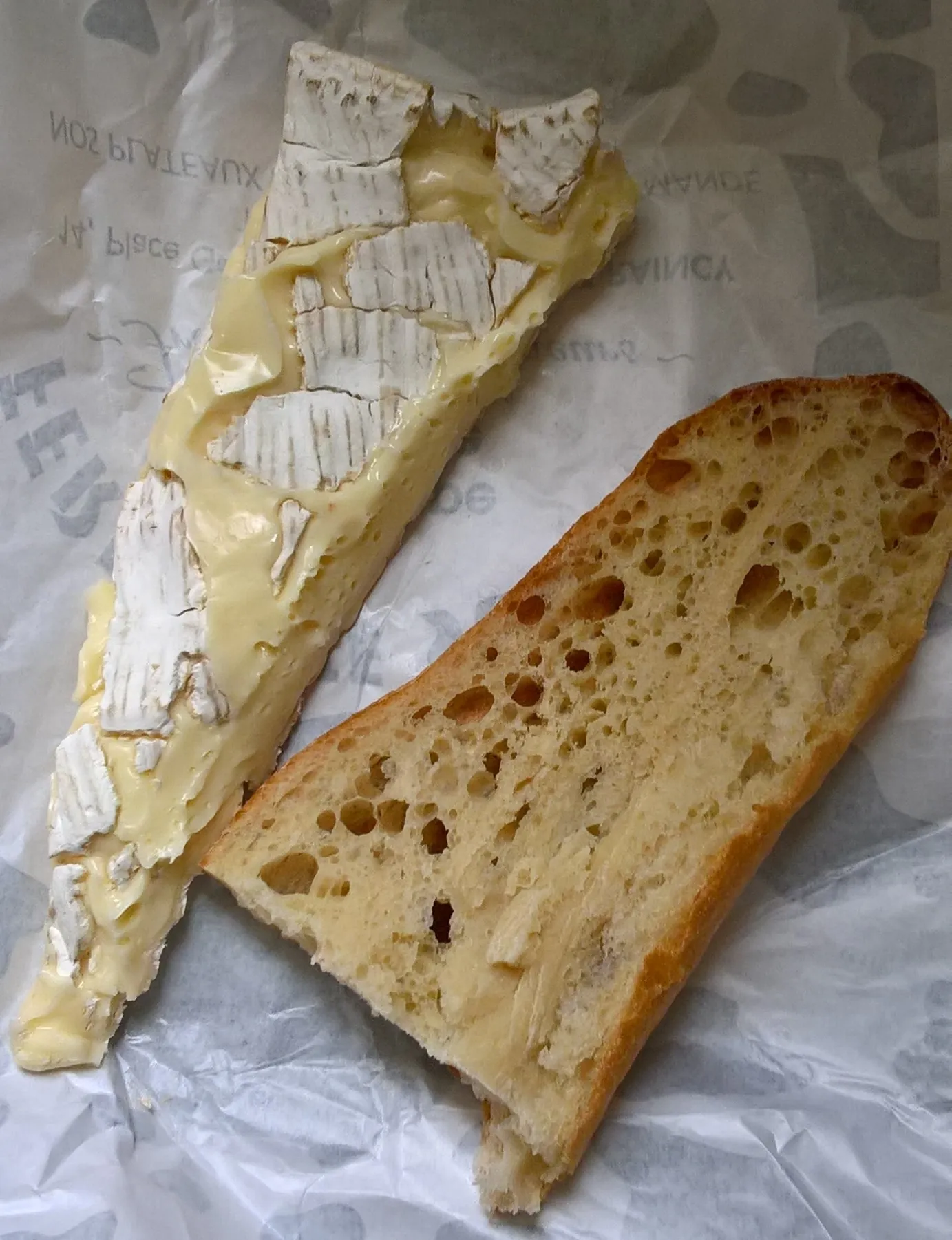 A long wedge of Brie de Meaux with its white rind beside a piece of crusty baguette on paper.