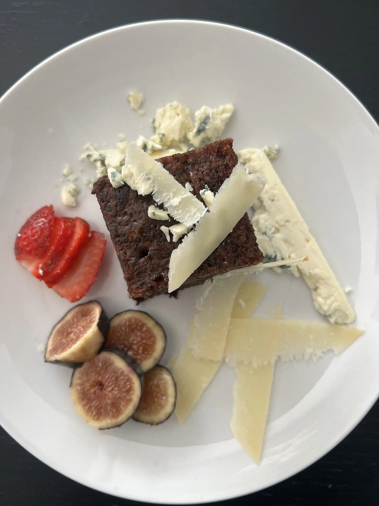 A white plate with a square of dark cake topped with blue cheese crumbles and shaved cheese, beside fresh figs and sliced strawberry.