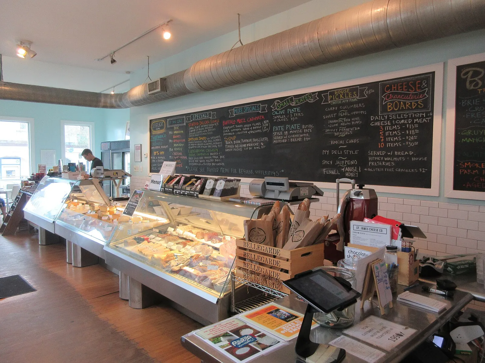 A cheese shop counter with a long glass case of cheeses, wrapped wedges in wooden crates and a large chalkboard menu on the wall behind