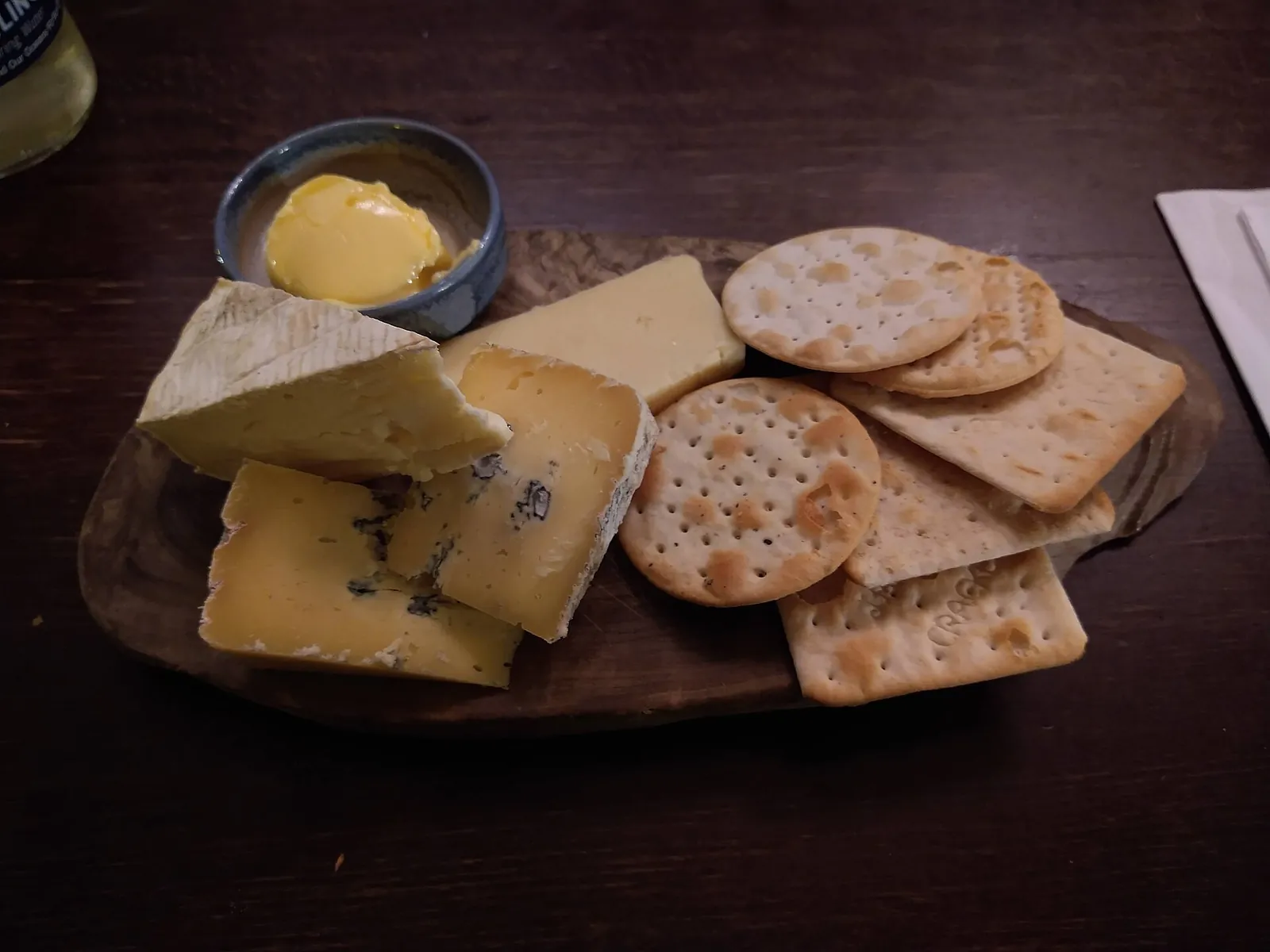 A dark wooden board holding wedges of soft white-rind cheese and blue-veined cheese, a small dish of butter, and round and square crackers.