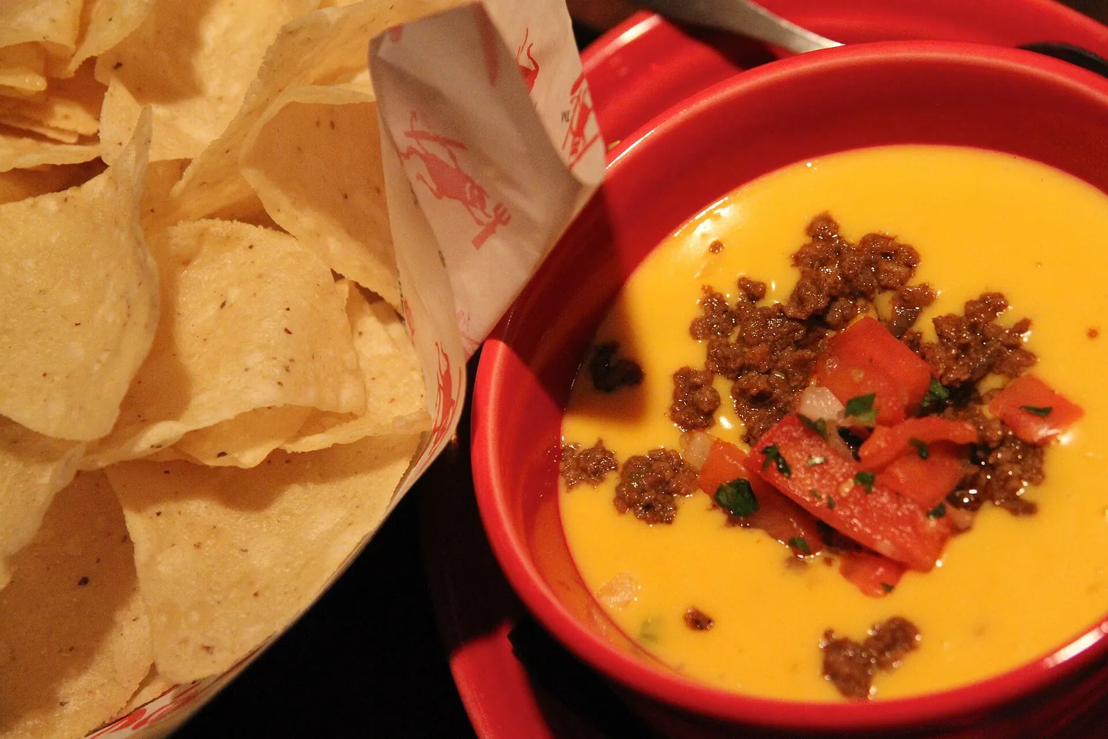 A red bowl of warm, bright orange chile con queso topped with chorizo and diced red pepper, with tortilla chips beside it.