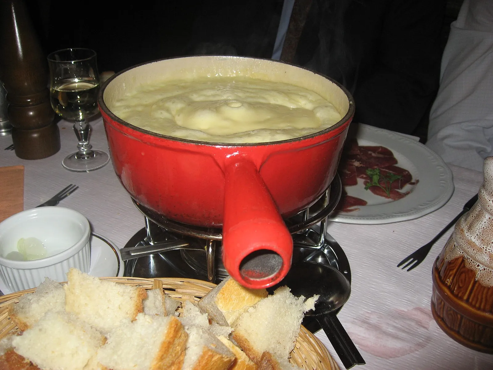 A red enameled fondue pot of smooth melted cheese over a burner on a table, with a basket of bread cubes in front.