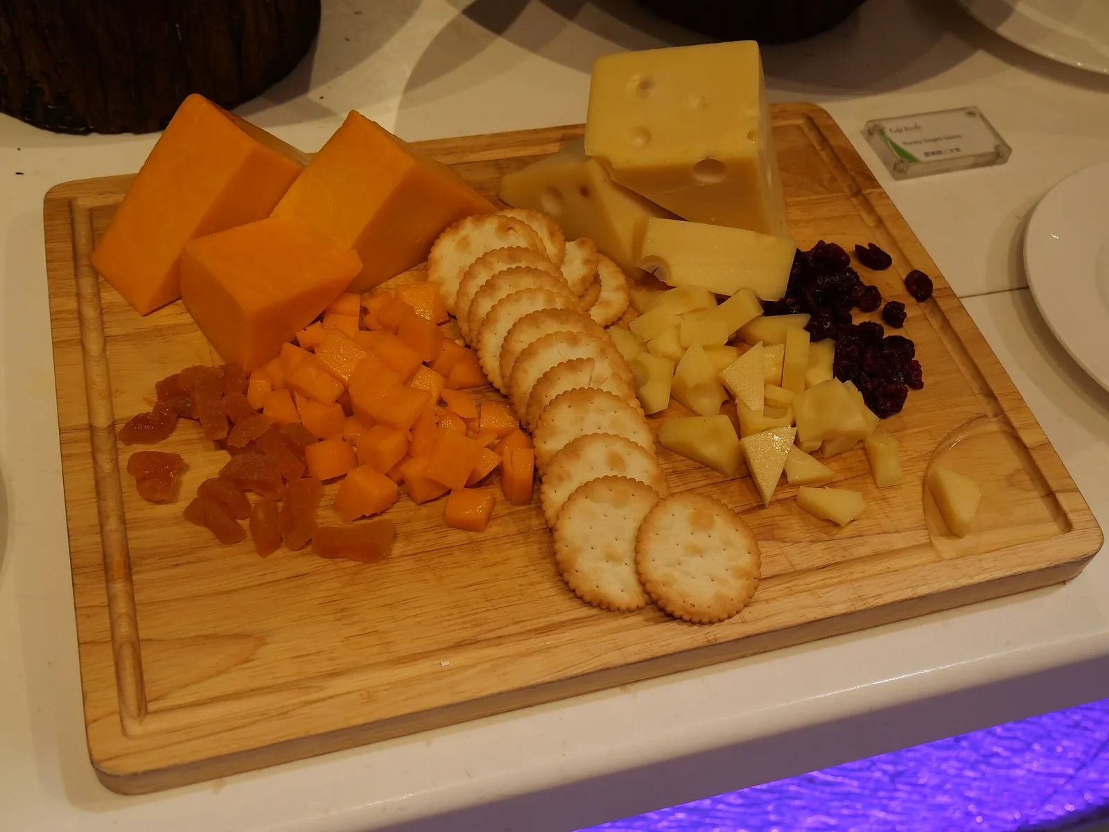 A wooden board with orange cheddar cubes, pale cheese slices, round crackers and dried fruit arranged as a snack plate.