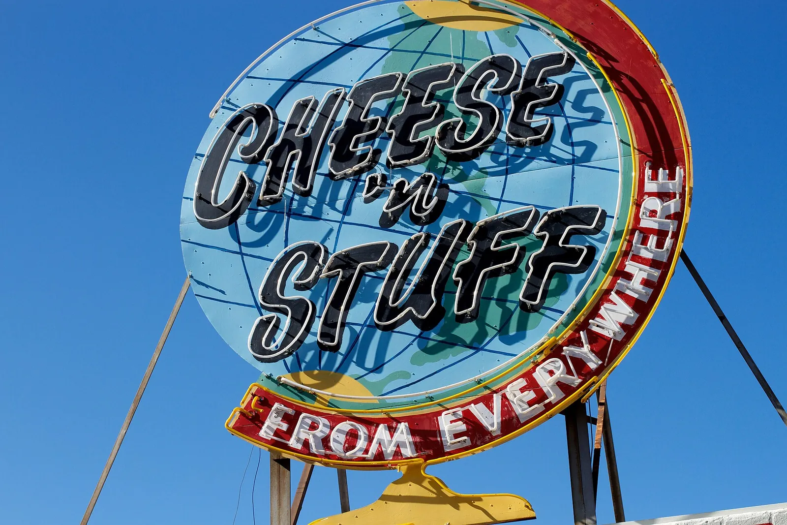 A round neon deli sign shaped like a globe, reading Cheese 'n Stuff From Everywhere, against a blue sky