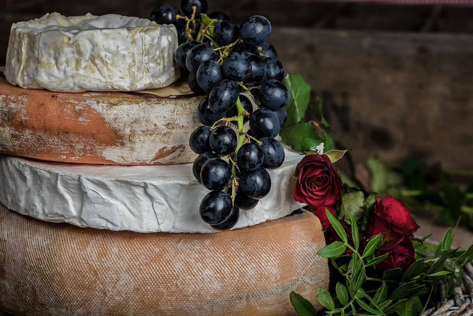 Stacked cheese wheels with white and orange rinds beside a bunch of dark grapes and red roses on a rustic table.