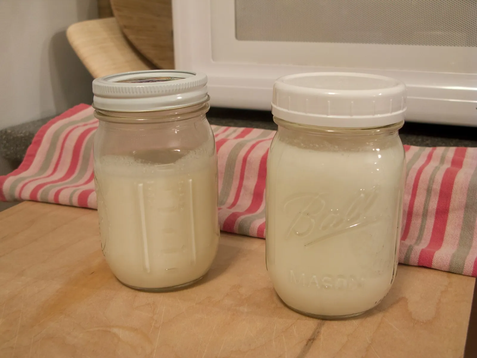 Two glass jars of creamy white kefir resting on a wooden cutting board in a kitchen.