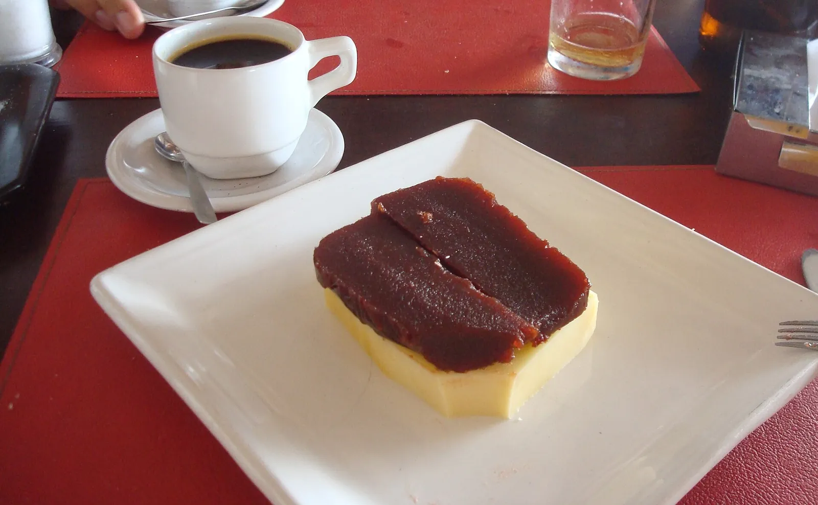 A thick slab of quince paste on a slice of pale cheese on a white square plate, with a cup of black coffee behind.