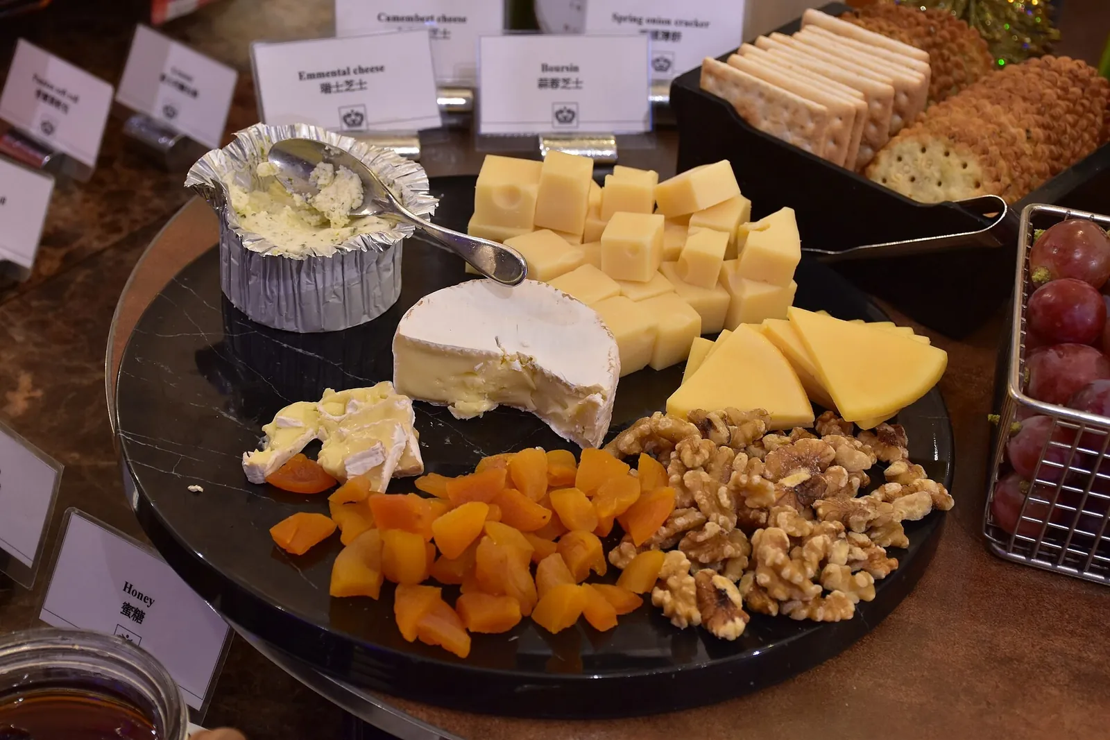 A round black platter of cheeses with cubes of yellow cheese, a soft white wedge, dried apricots and a pile of walnuts.