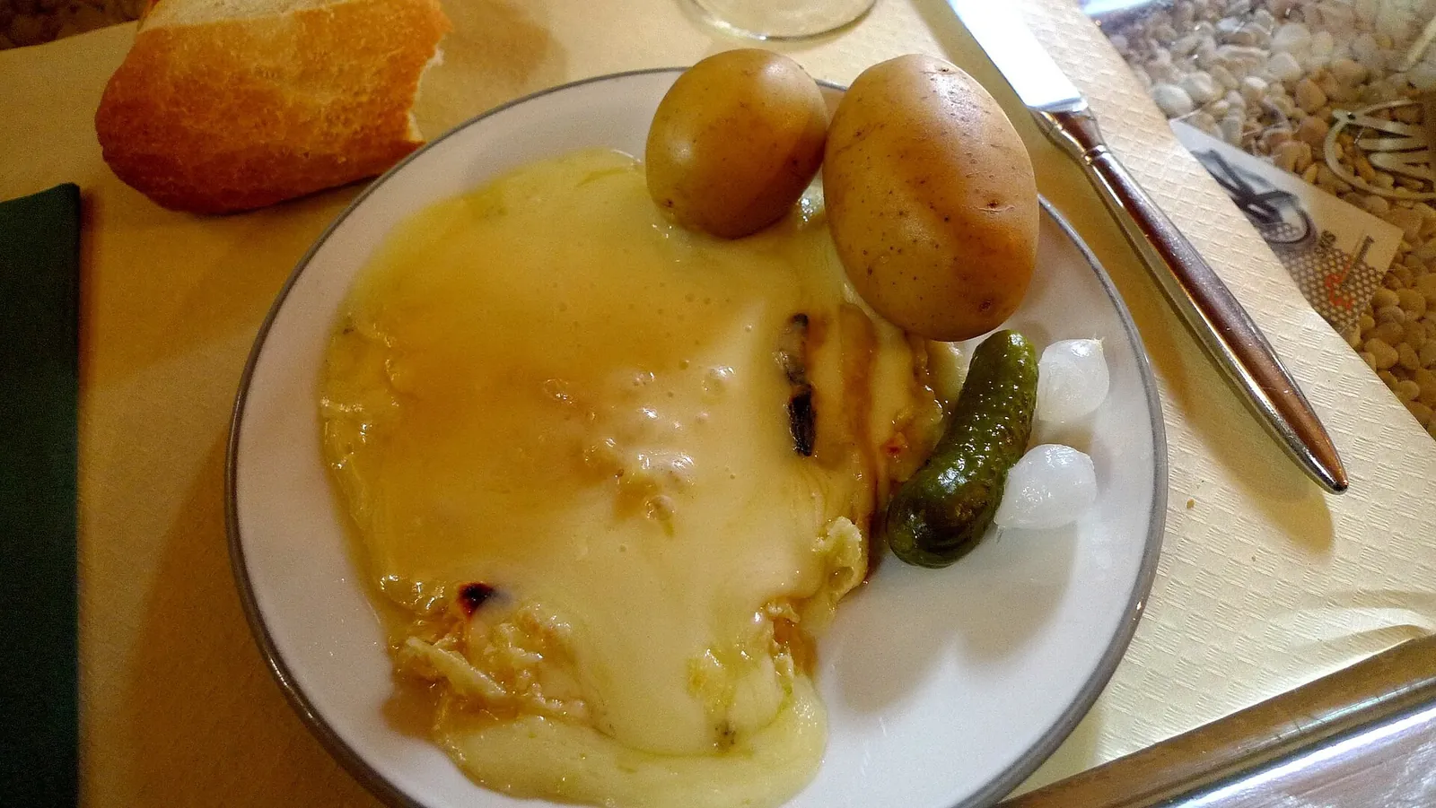 A white plate of melted raclette cheese with two boiled potatoes, a cornichon and pickled pearl onions, with bread and a knife beside it.