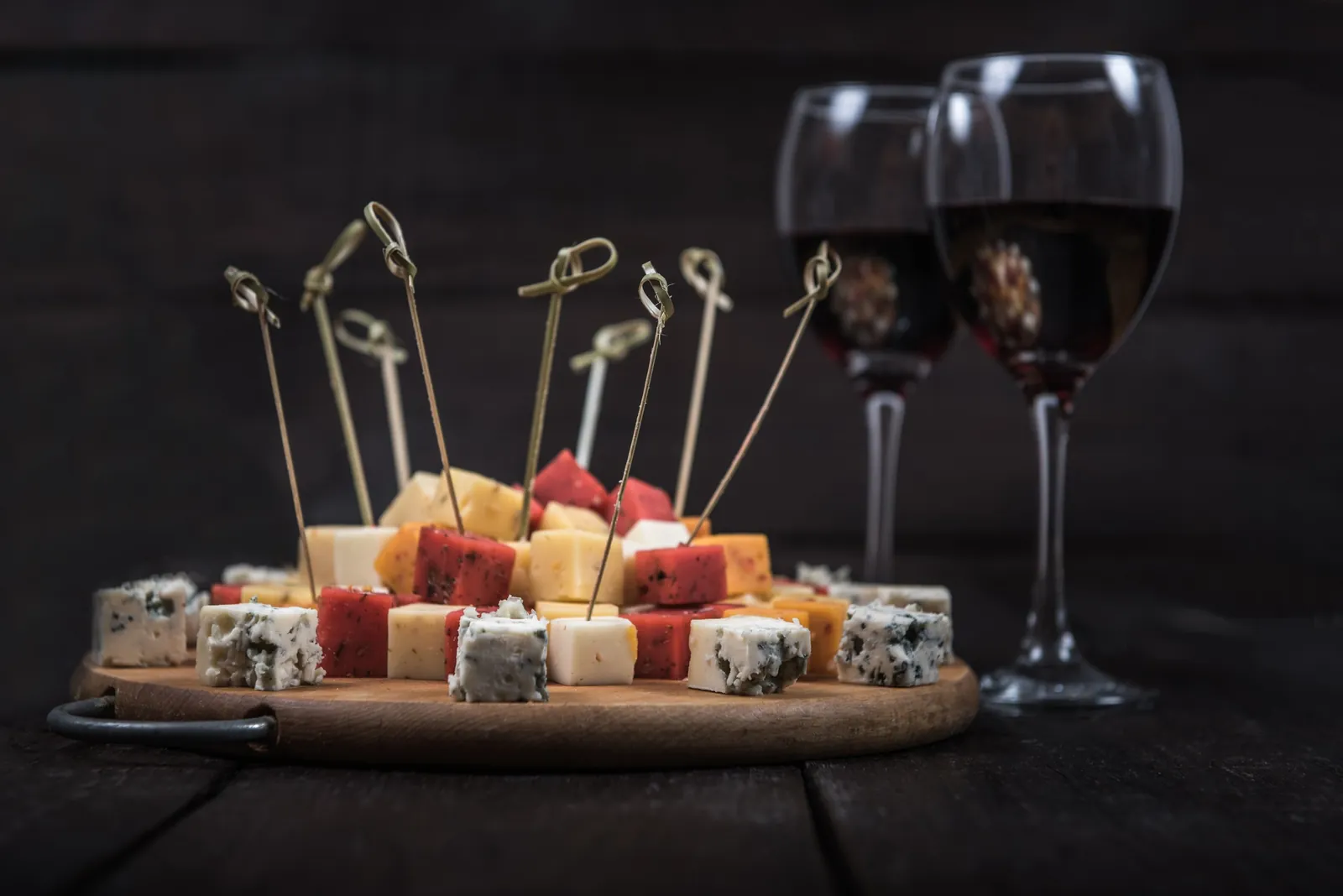 A wooden board of bite-size cheese cubes on bamboo picks, including blue and red-rind cheeses, with two glasses of red wine behind.
