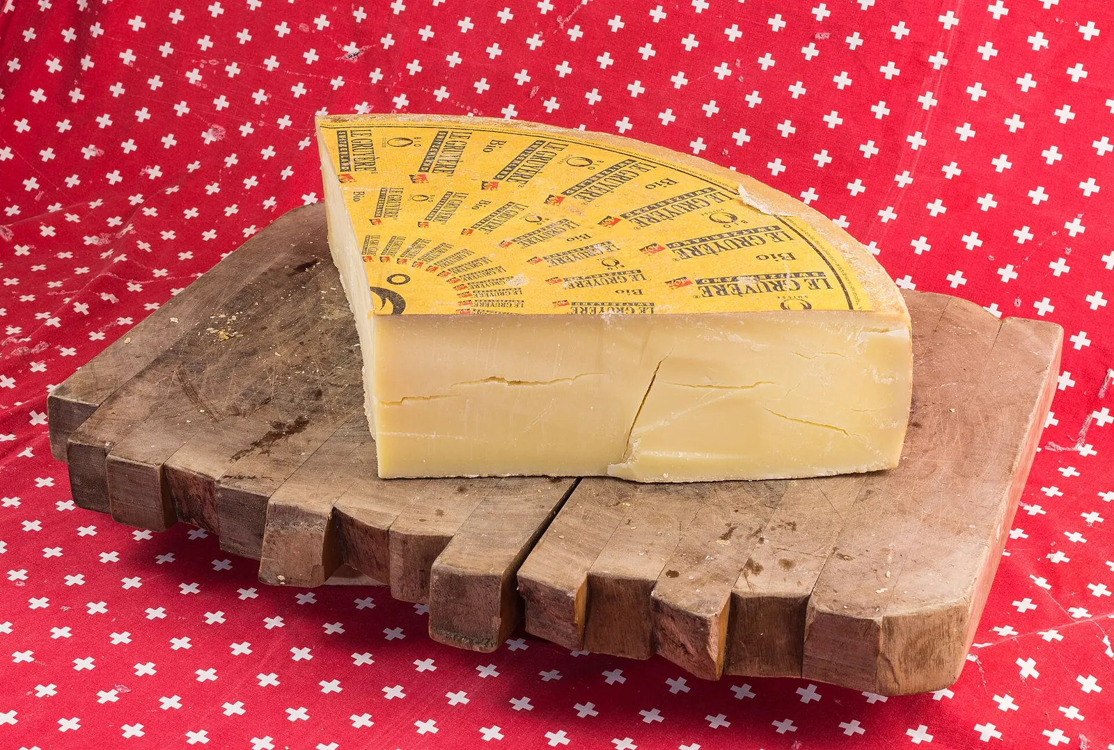 A large wedge of hard Gruyère cheese with its rind still on, resting on a rustic wooden board over a red dotted cloth.
