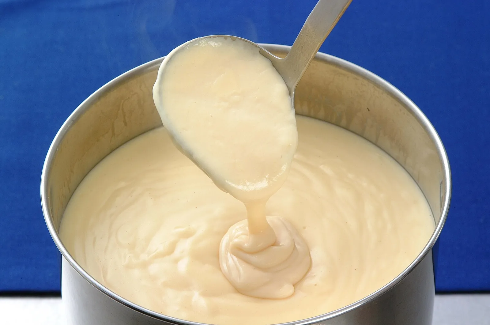 A ladle lifting a thick, smooth white cheese sauce from a steel pot, with a ribbon of sauce falling back in.