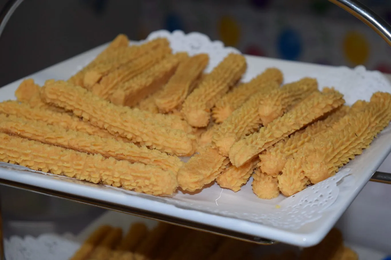 A white tray lined with a paper doily holding golden, ridged cheese straws piped and baked into long twists