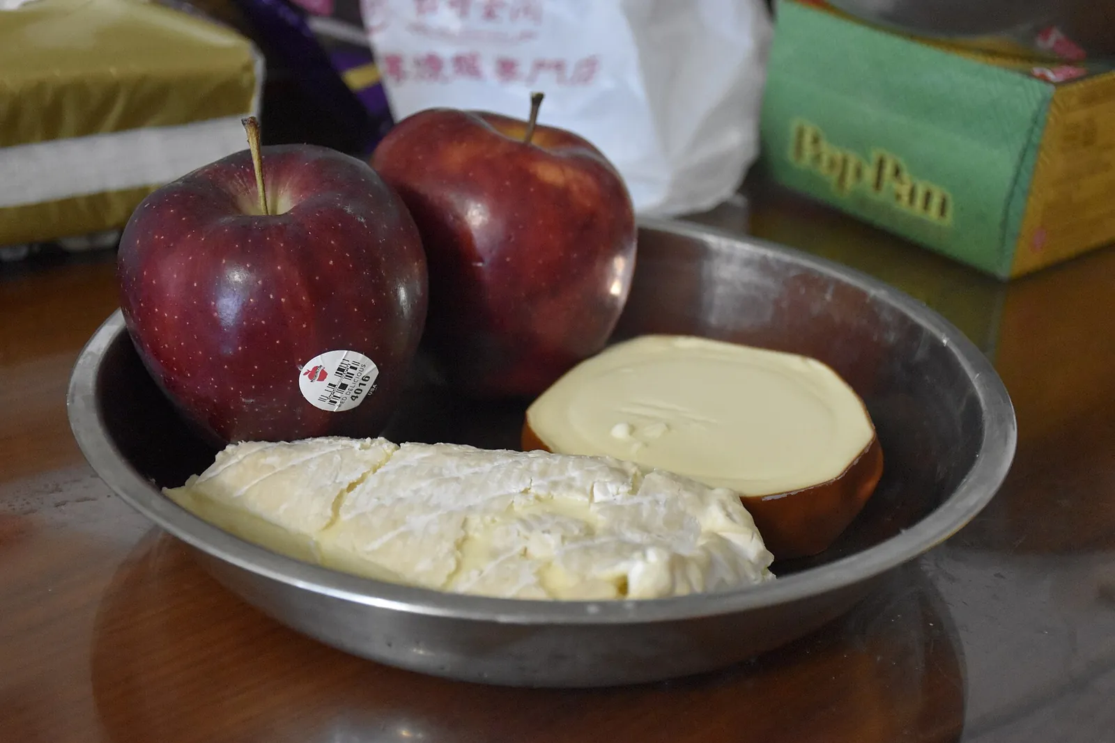 Two red apples in a metal bowl beside a wedge of soft white cheese and a piece of hard cheese.