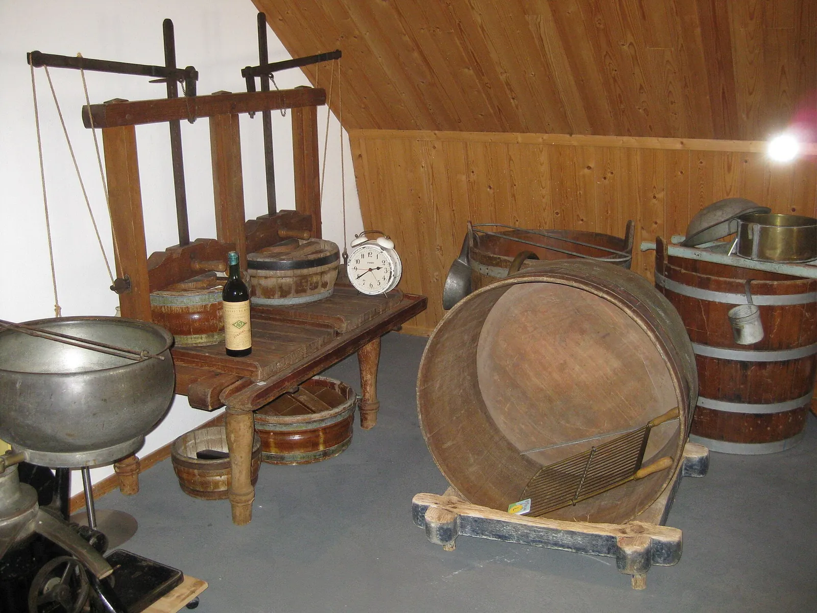 An old cheesemaking workshop display with a wooden cheese press frame, a large round sieve, wooden tubs and a steel cheese pot.