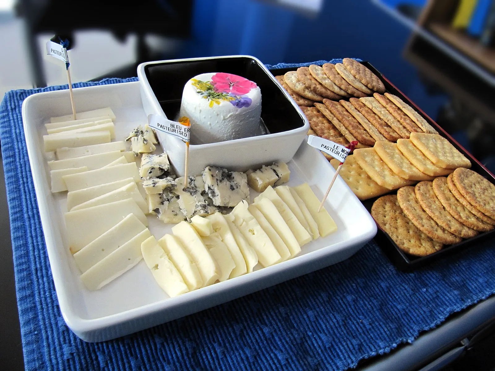 A white platter of sliced cheeses, blue cheese and a small round topped with edible flowers, beside a tray of crackers