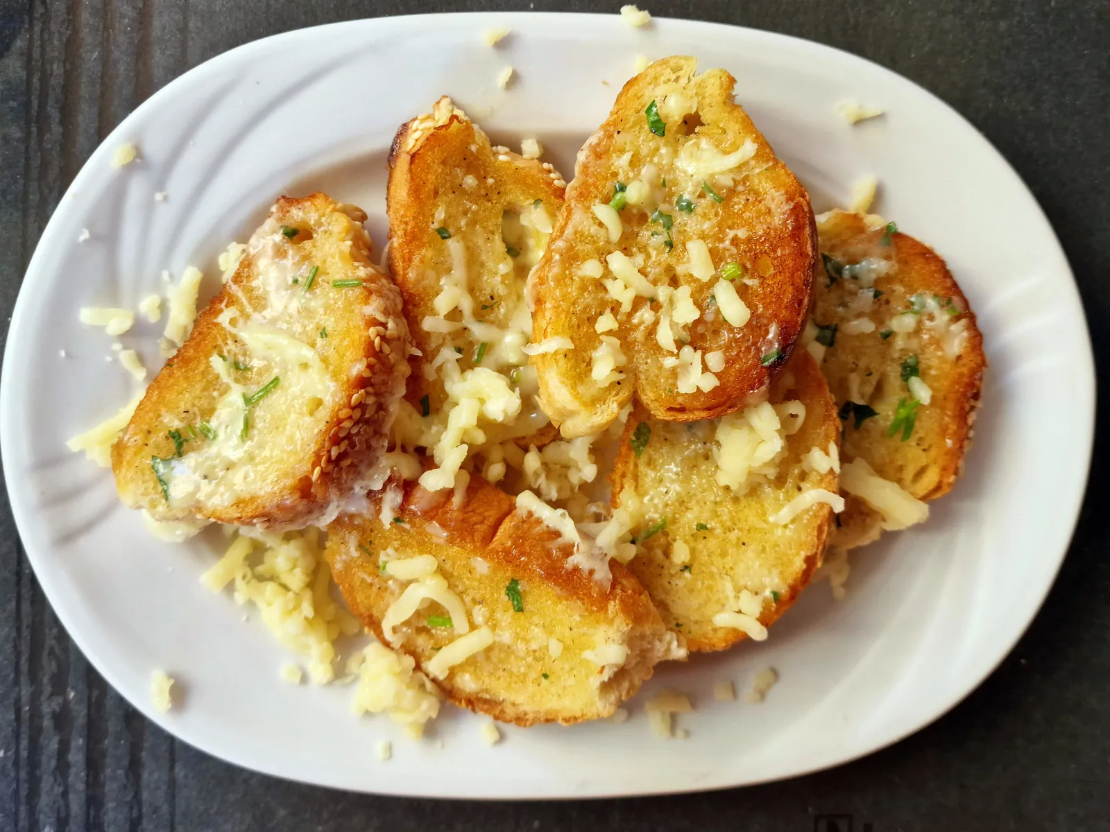 Slices of toasted garlic bread topped with melted, browned shredded cheese and herbs, piled on a white plate