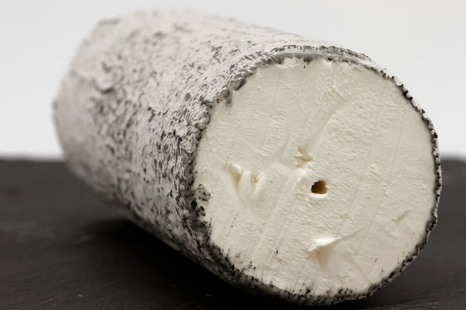 A log of Sainte-Maure de Touraine goat cheese with a gray ash rind, cut to show the bright white paste
