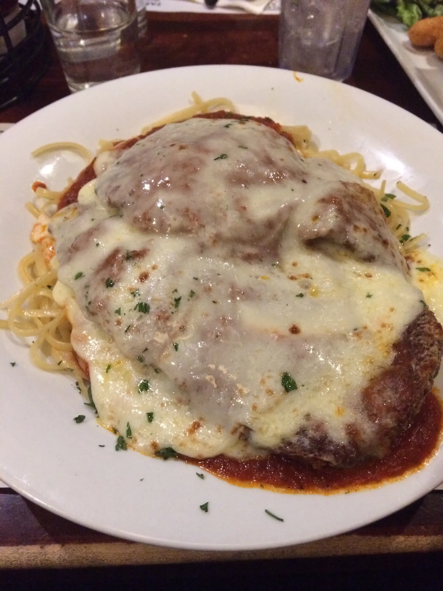 A breaded chicken cutlet covered with melted mozzarella and tomato sauce, served on a plate over spaghetti and sprinkled with parsley.