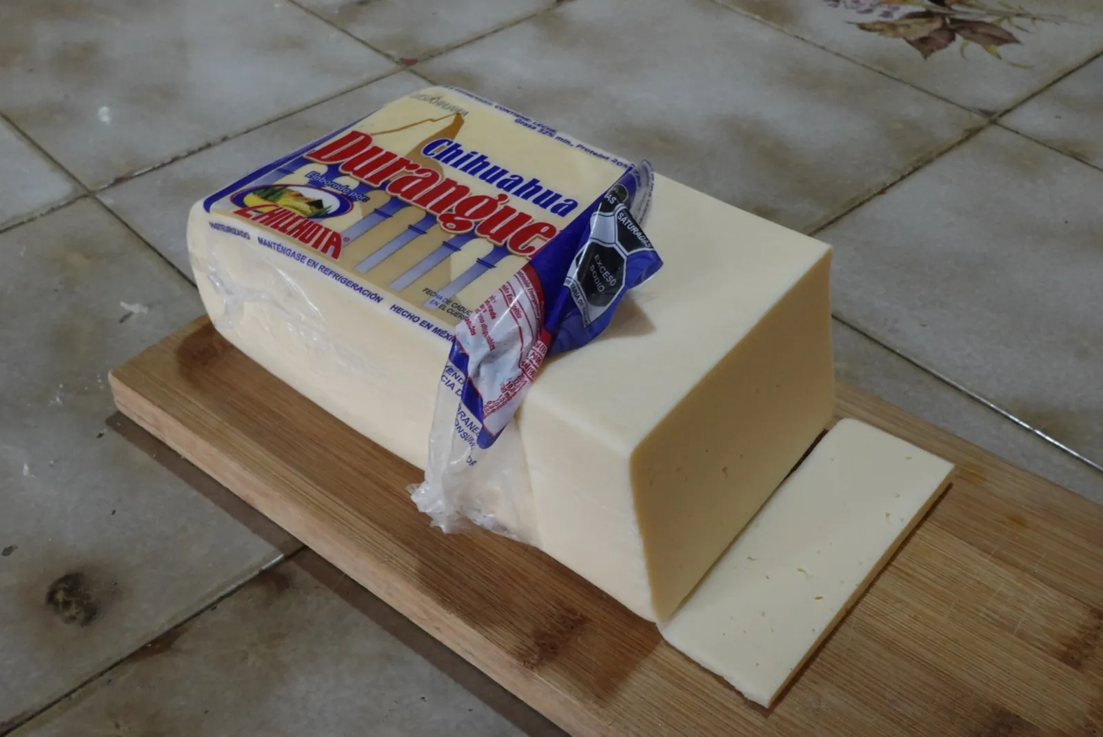 A block of Chihuahua cheese partly unwrapped from its package with one slice cut off, on a wooden board